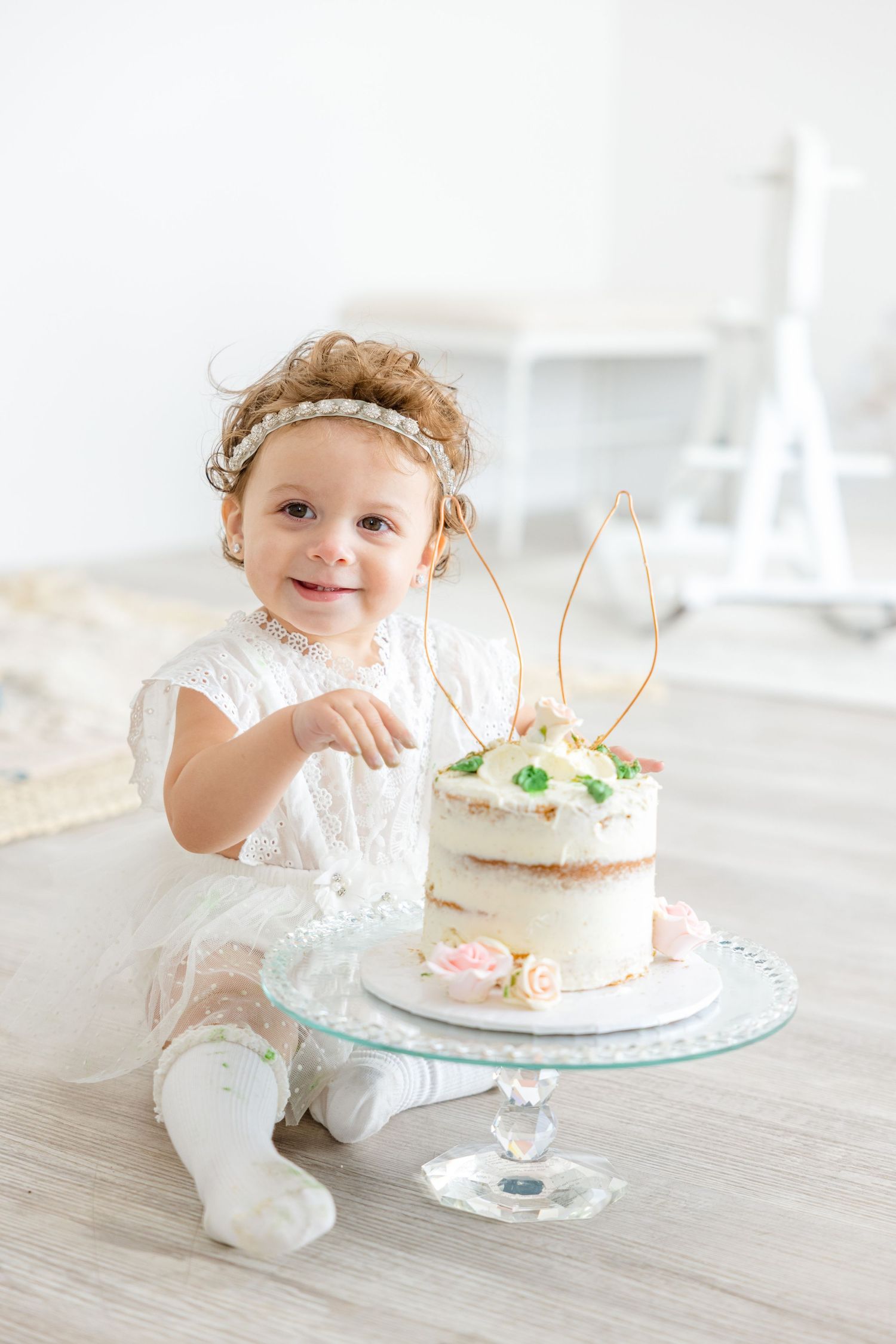 A toddler in white dress points excitedly at a decorated cake with wire bunny ears on top.
