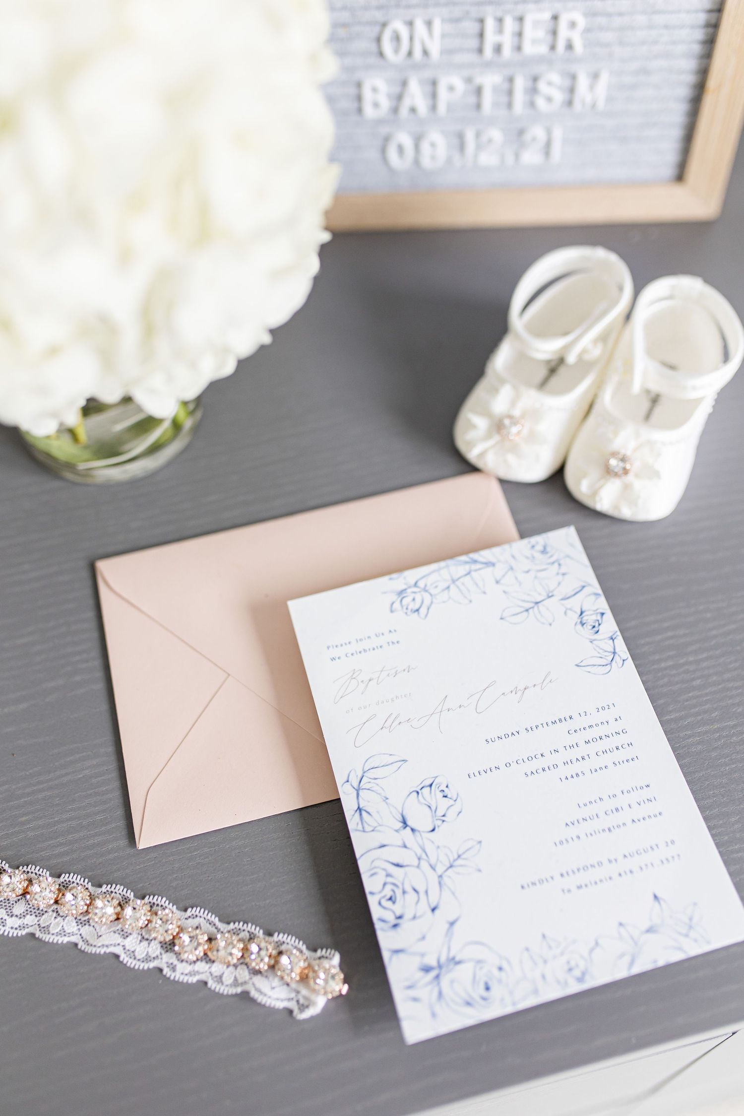 Baptism invitation with pink envelope next to white shoes and letterboard on grey surface.
