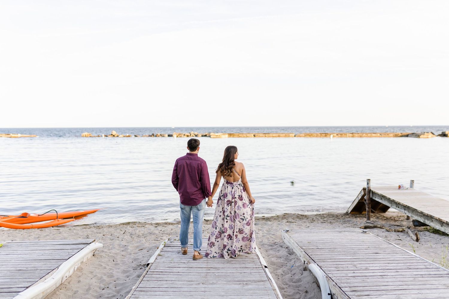 Couple walking away on wooden pier with water and boats in background.