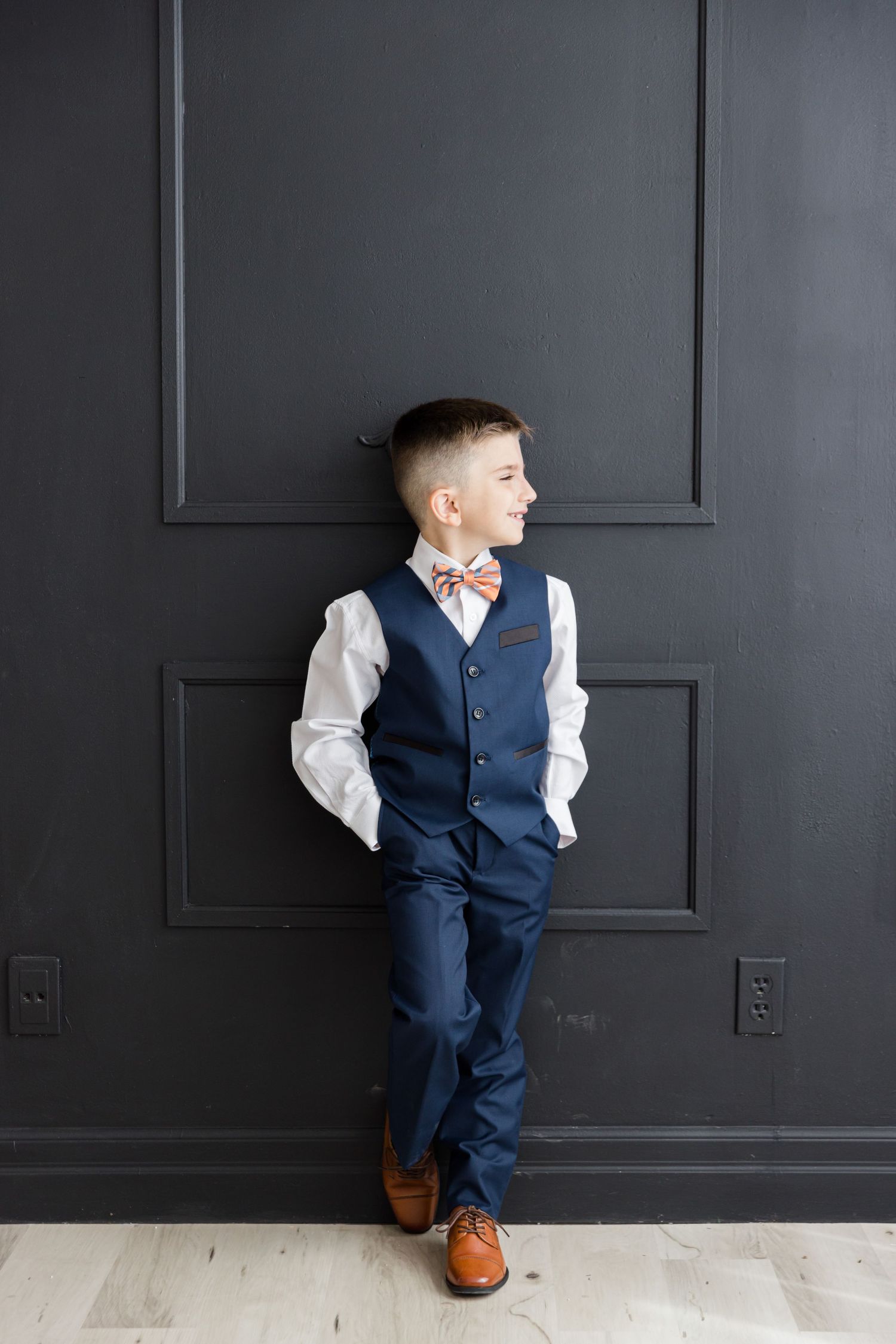 Side profile pose against dark wall wearing navy vest and brown leather shoes with hands in pockets.