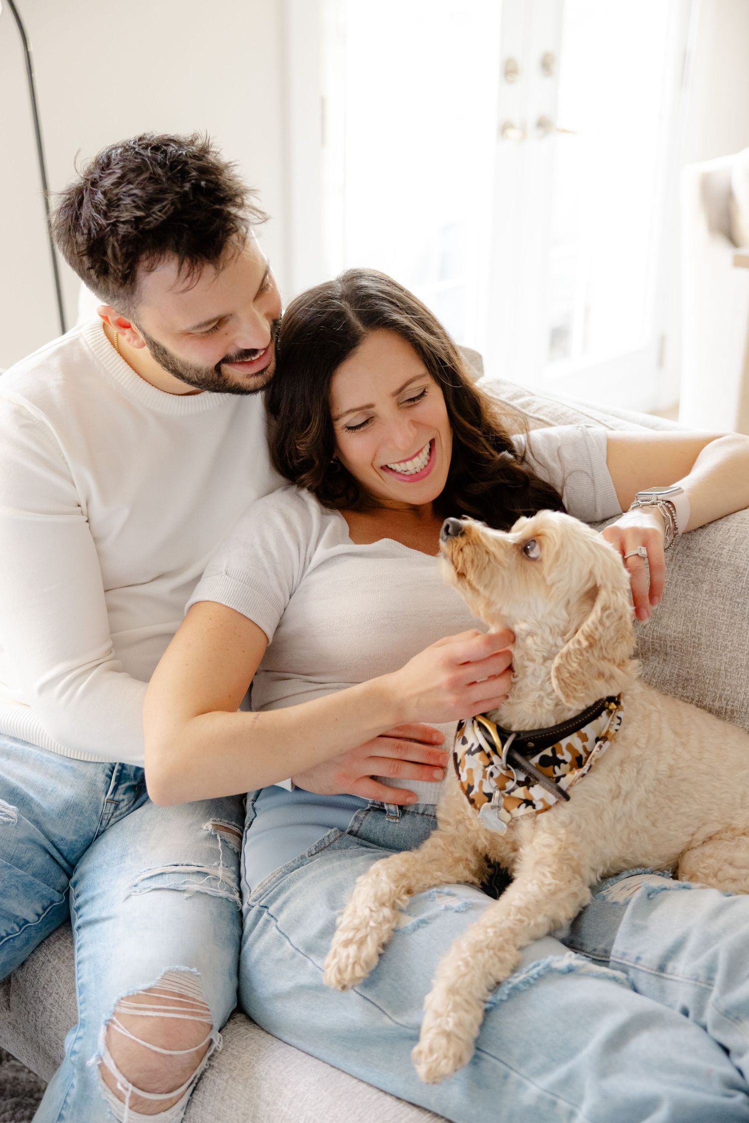 A couple on a couch cuddles with their fluffy dog, sharing affectionate moments in a bright, cozy living room.