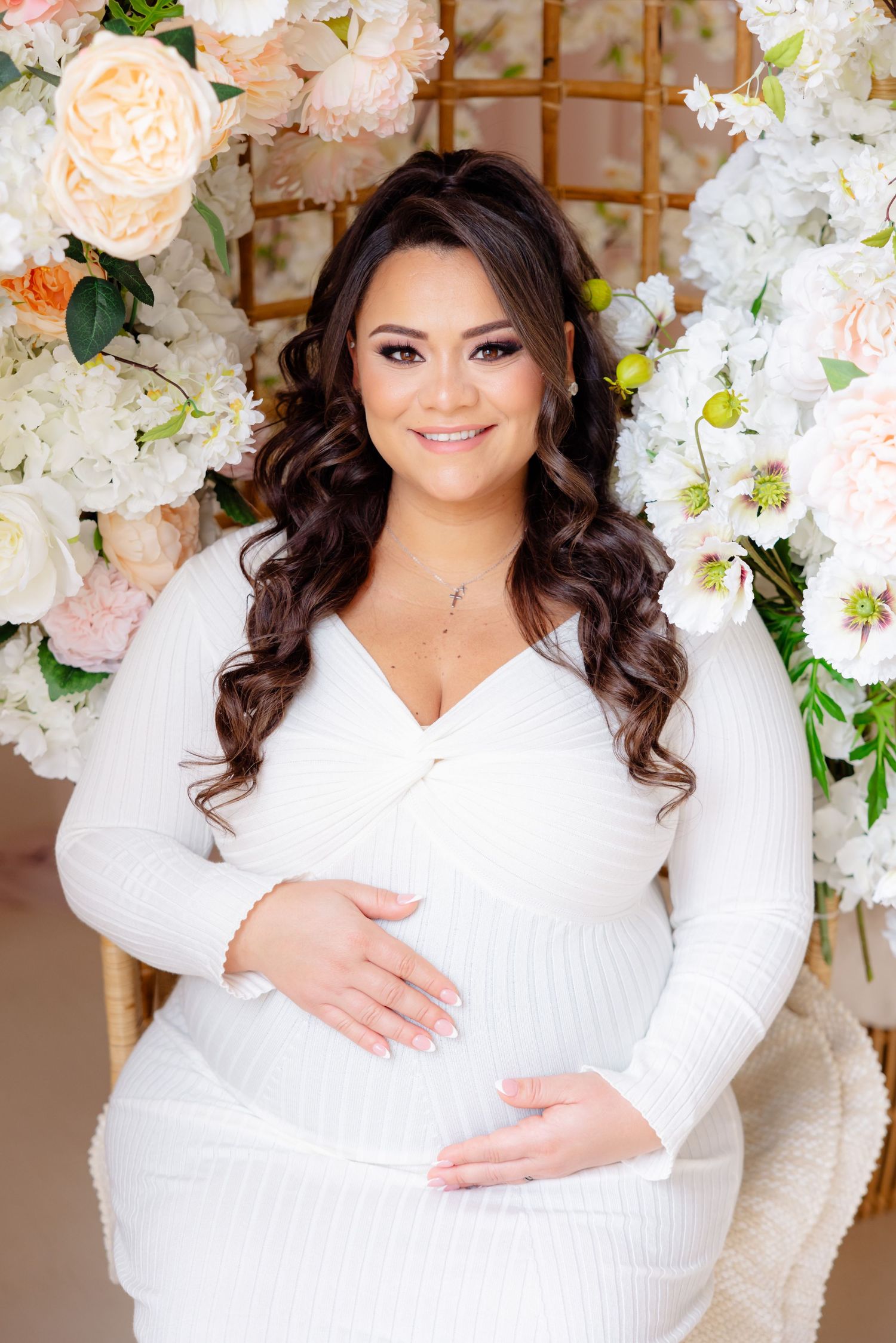 An expectant mother in a white dress poses in front of a floral backdrop with pink and white flowers.