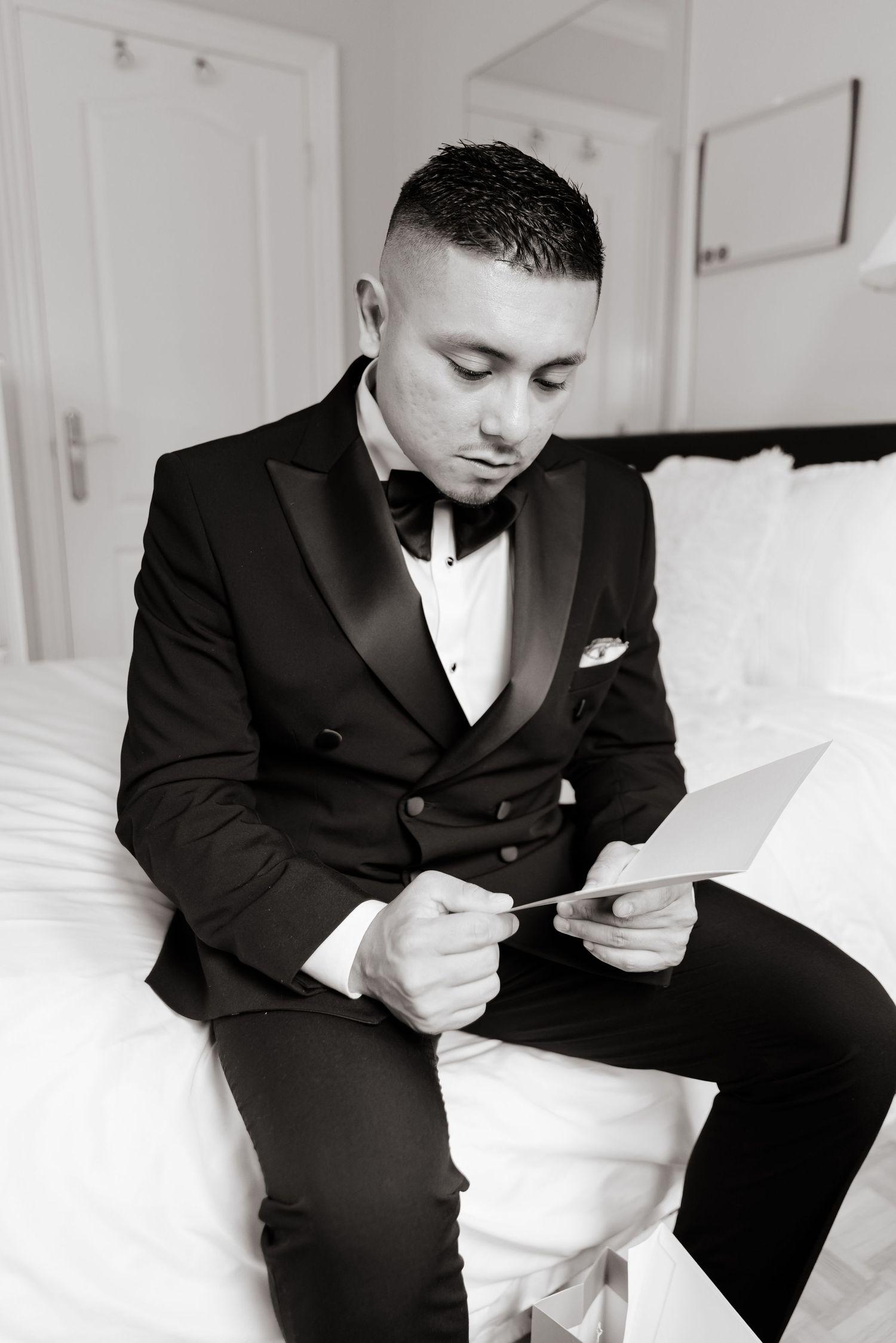 A black and white photo shows someone in a formal tuxedo reading a note while sitting on a white bed.