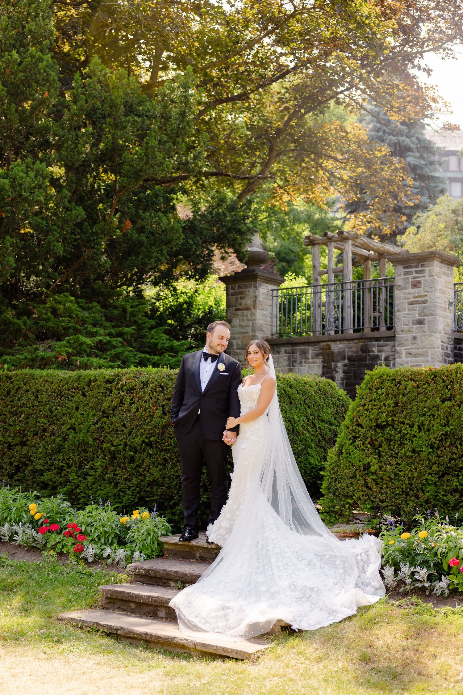 Bride in flowing white gown and groom stand on garden steps surrounded by lush greenery and stone architecture.