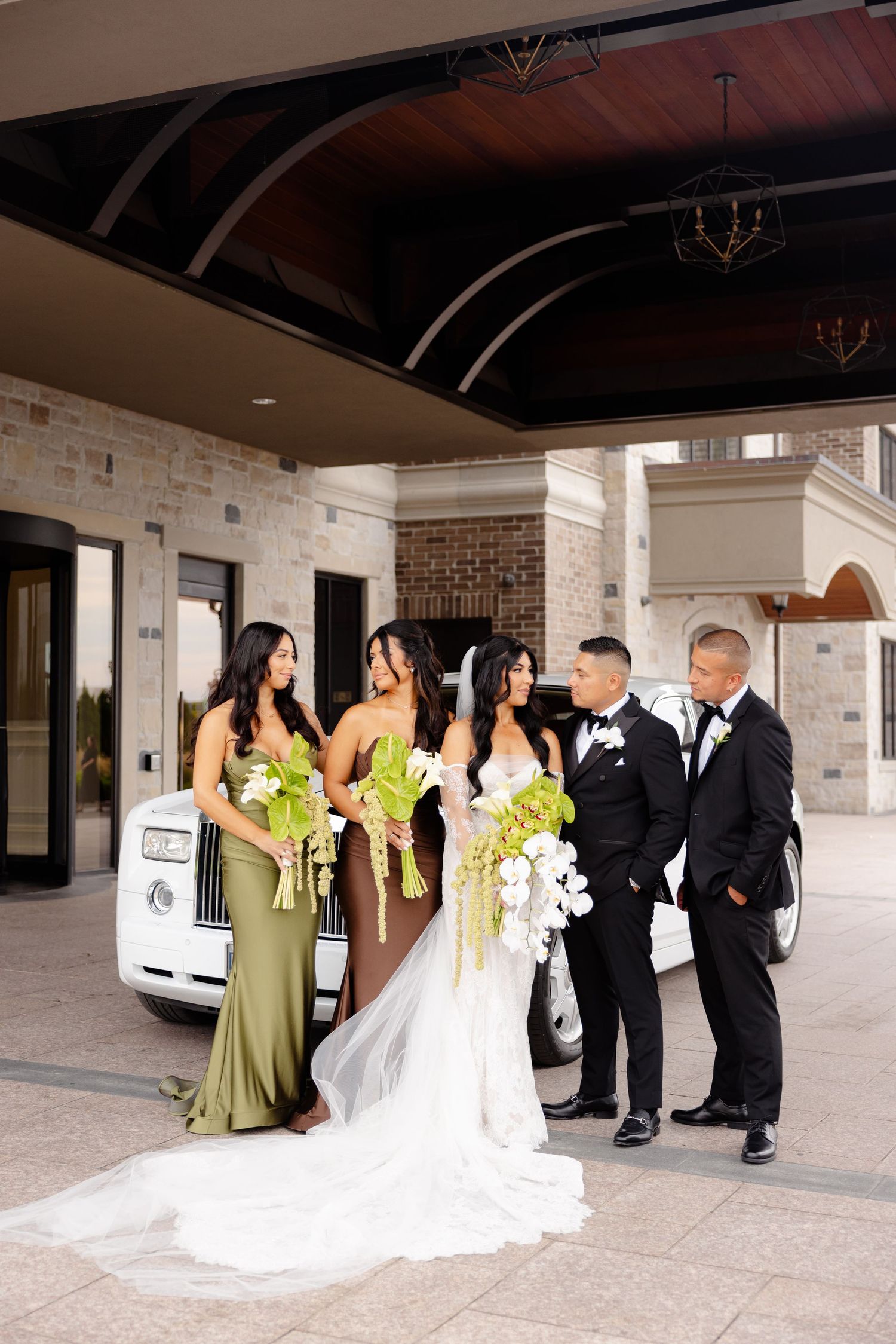Wedding party dressed in black and olive green poses together under covered entrance of brick building for formal photos.