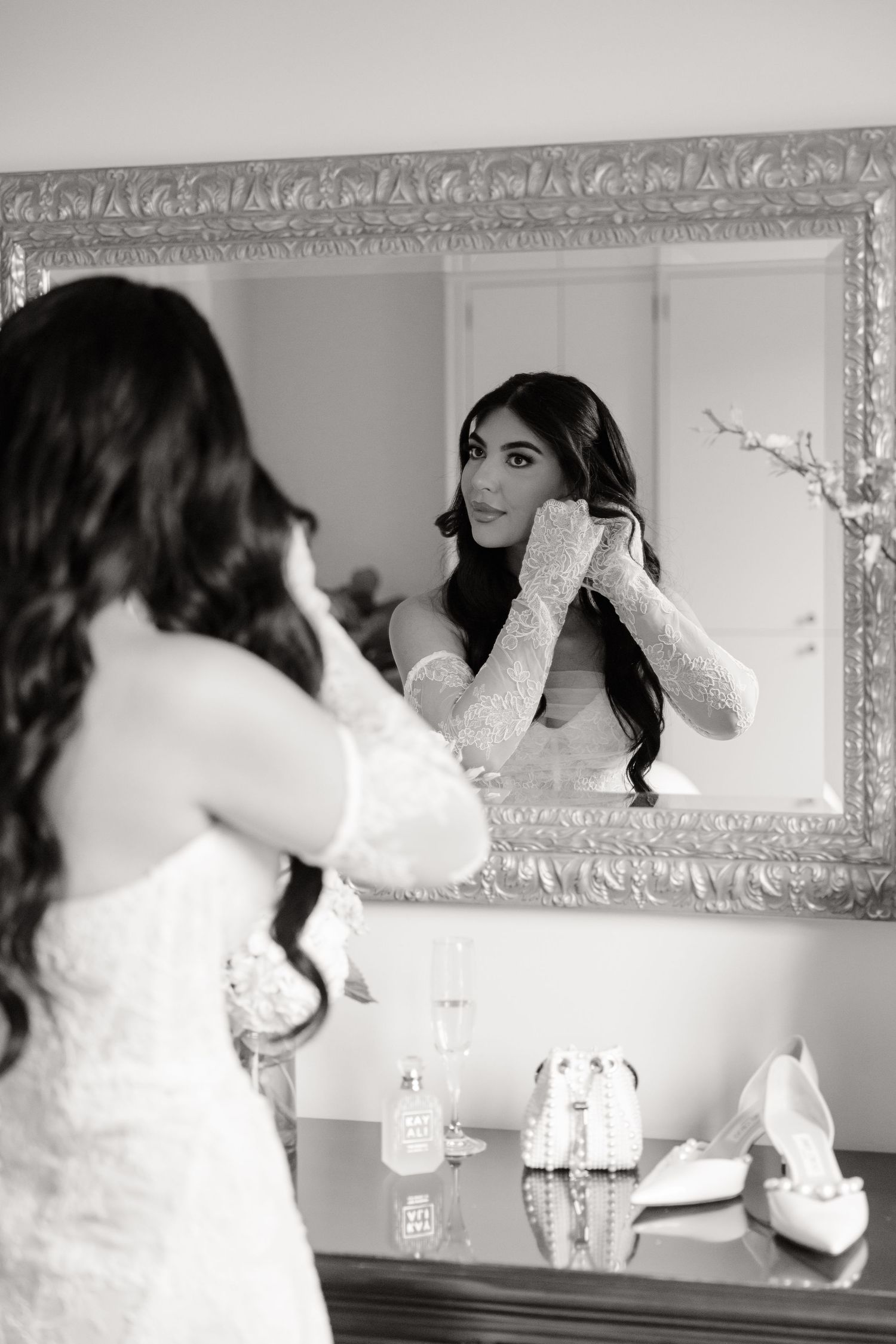 A reflection in an ornate silver mirror shows someone in a white lace wedding dress adjusting earrings before the ceremony.
