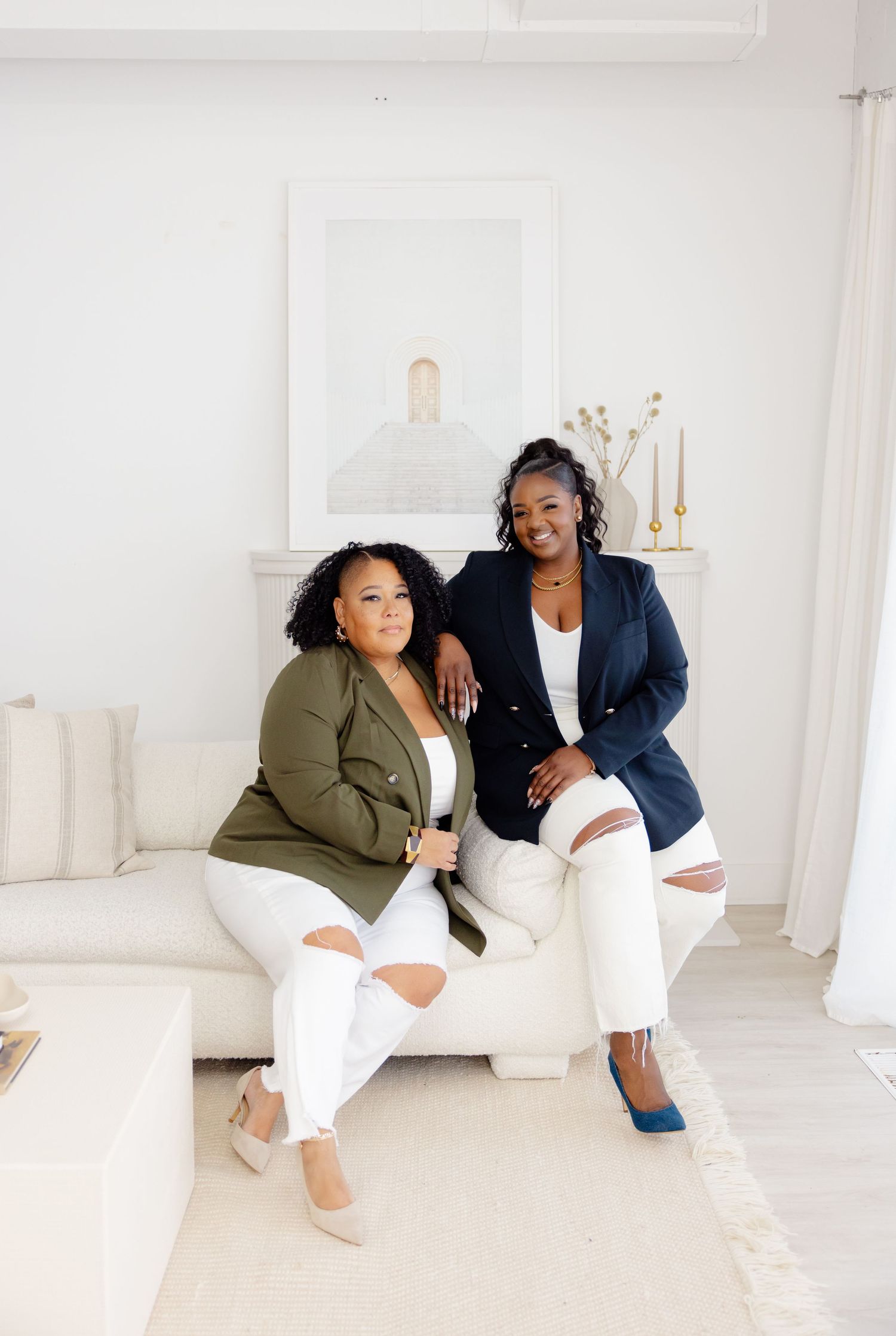 Two women in business attire pose together on white couch in a bright minimalist room with white decor.