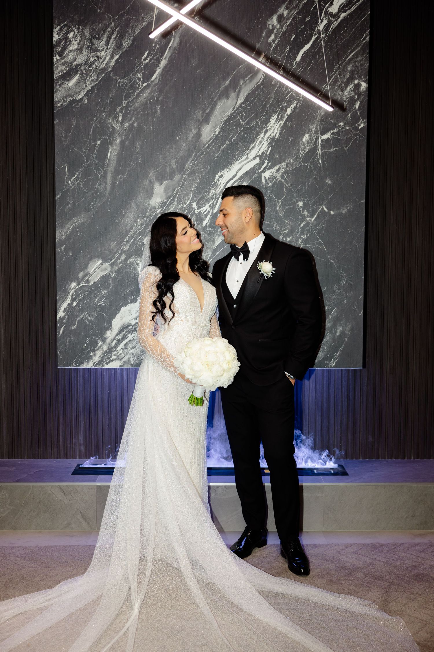 Romantic wedding portrait of couple against marble wall with geometric lighting fixtures.