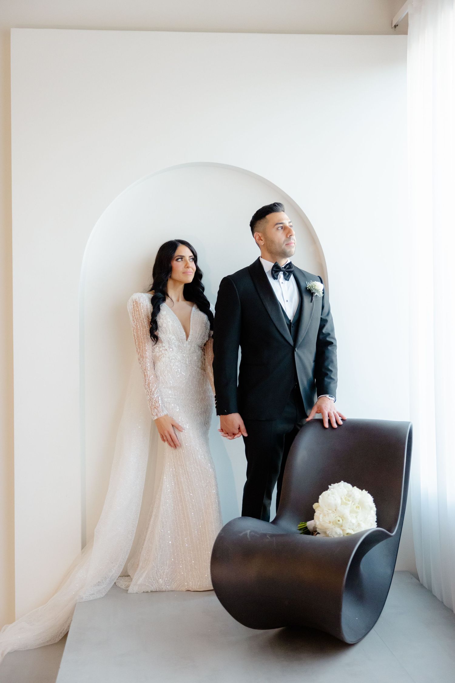 Elegant couple in wedding attire posed in minimalist white room with modern curved chair.