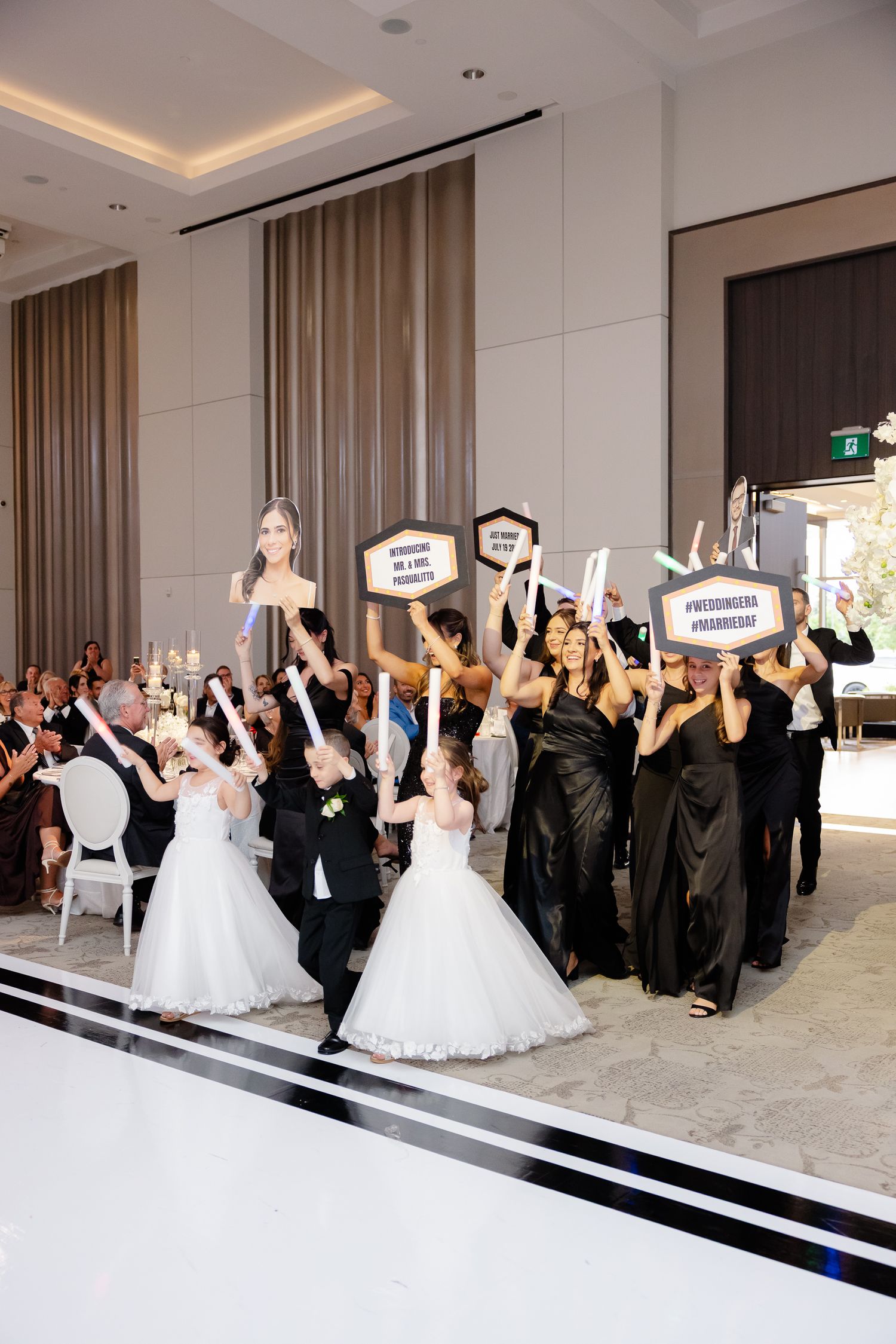 Wedding celebration with guests in formal attire holding up celebratory signs at an elegant hotel ballroom reception.
