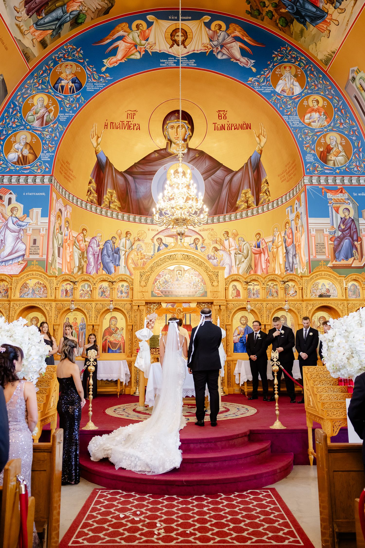 A wedding ceremony takes place inside an ornate Orthodox Christian church with golden Byzantine-style iconography and frescoes.