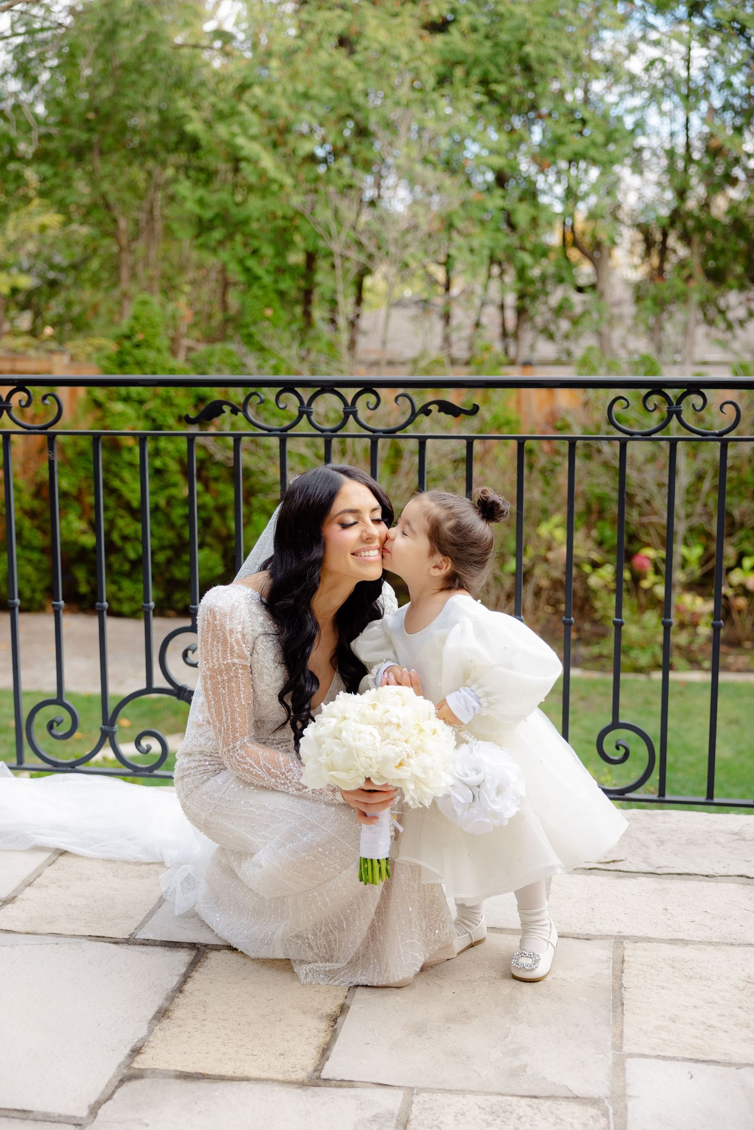 Touching candid moment between bride and flower girl on stone balcony with wrought iron railing.