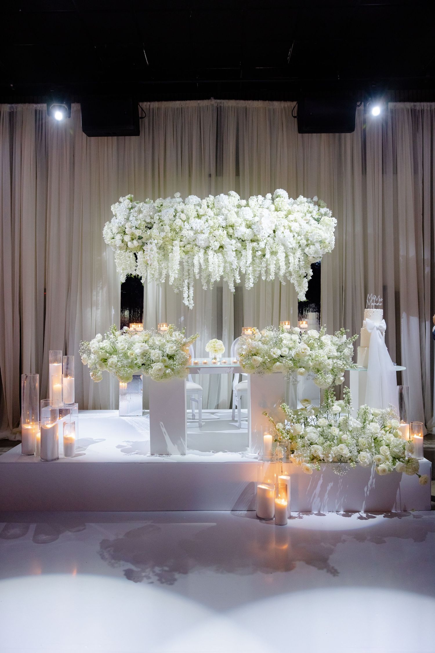 Dramatic white floral chandelier installation over wedding ceremony space with candlelit aisle decorations.