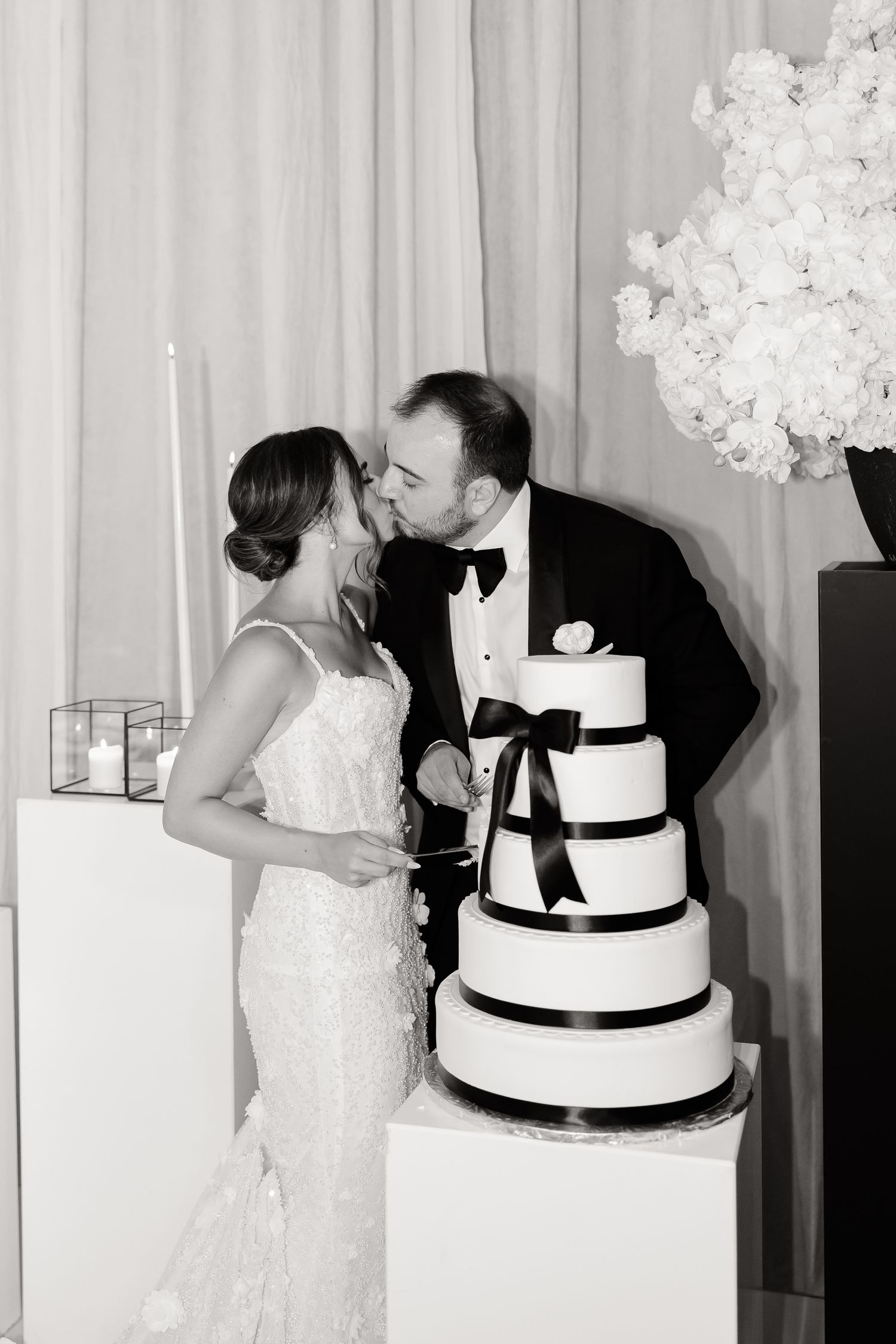 Elegant wedding moment with couple sharing a kiss next to their black and white tiered cake adorned with ribbons.