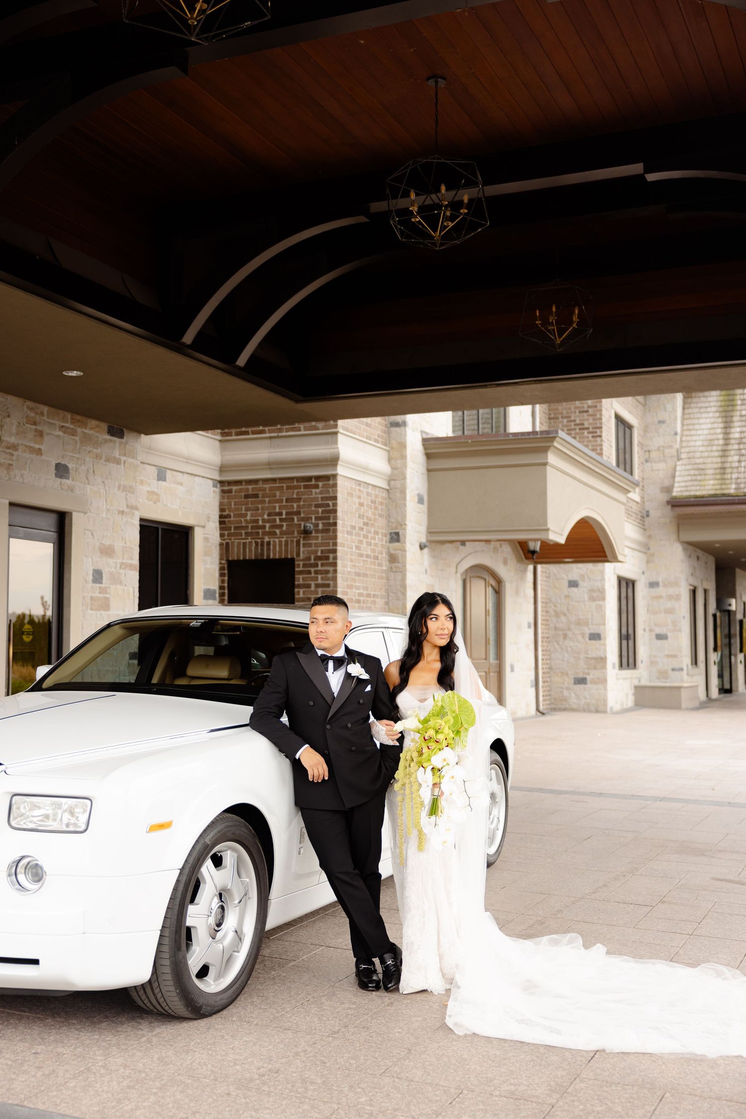 A wedding couple poses with an elegant white luxury car in front of a stone building with arched entryways.