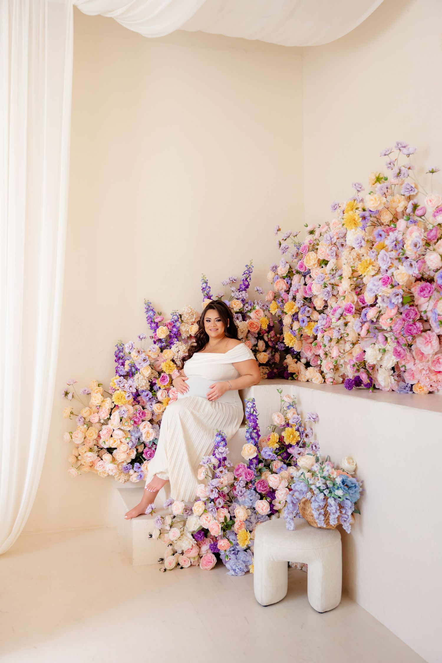 A dreamy maternity portrait surrounded by lush pink, purple and yellow floral arrangements in a light studio setting.