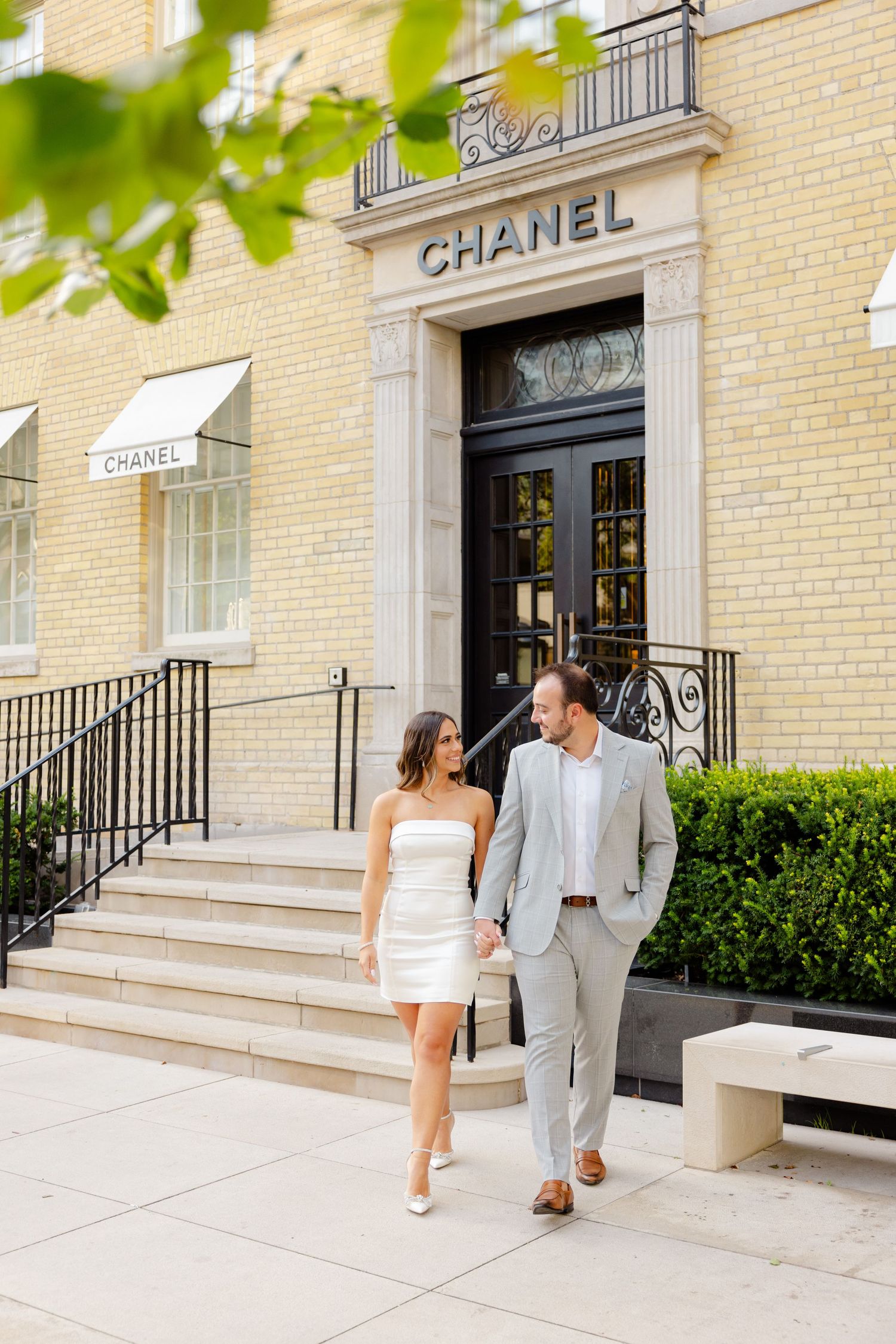 Couple walking down steps of luxury Chanel store with cream brick exterior and iron railings.