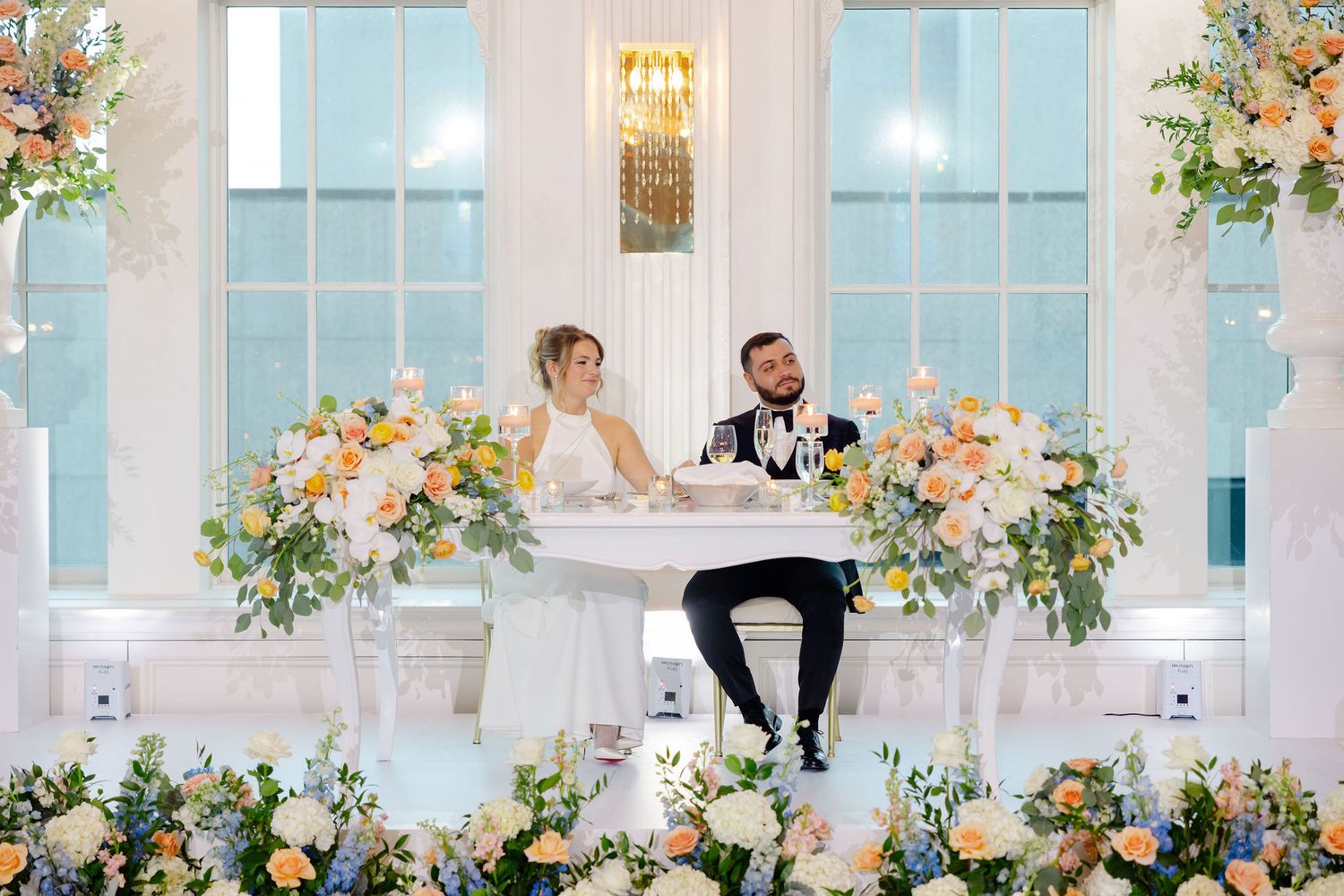 Elegant wedding reception setup with couple at sweetheart table adorned with lush floral arrangements and tall windows.