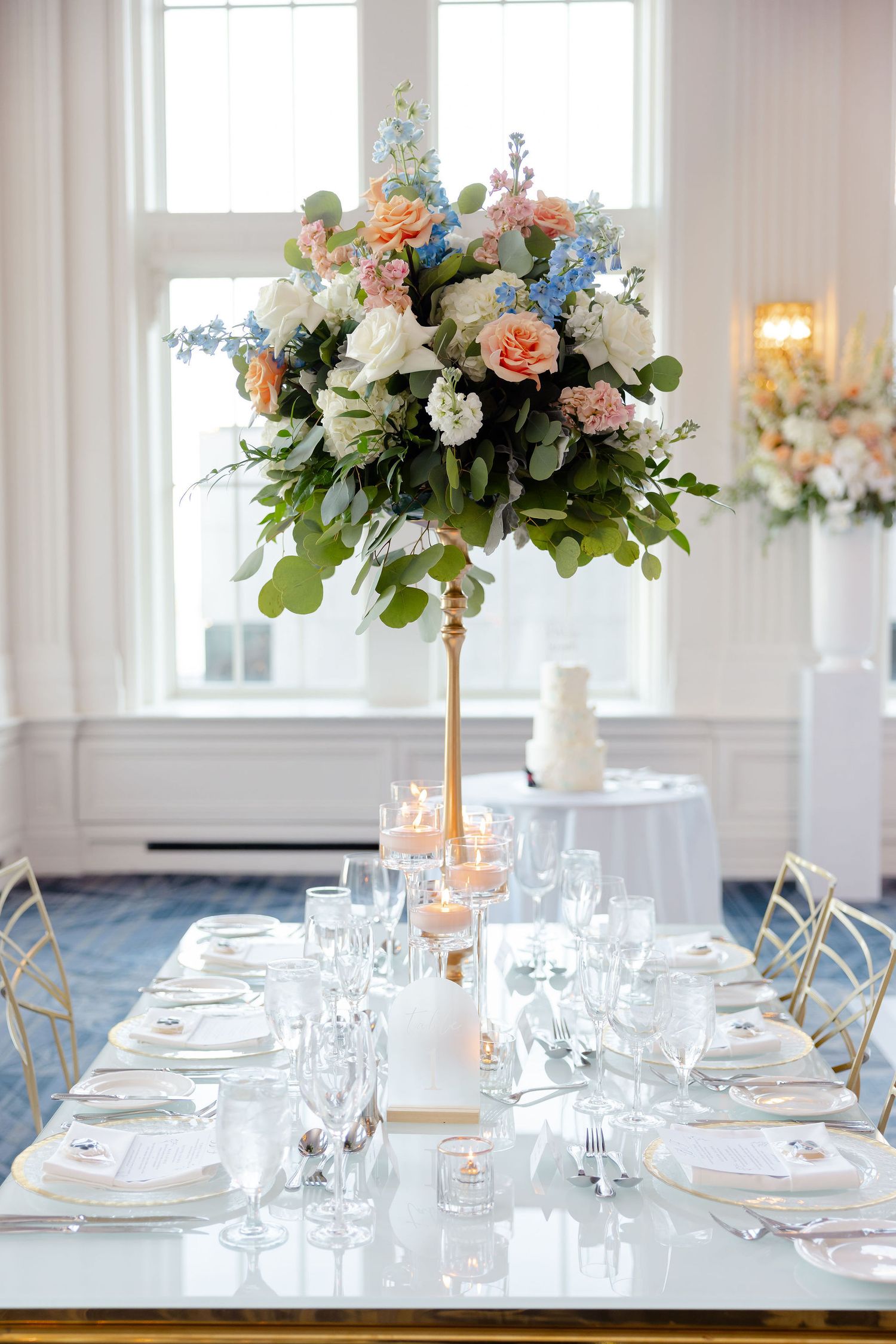 Elegant wedding table setting with tall floral centerpiece featuring peach roses and blue delphinium on gold stand.