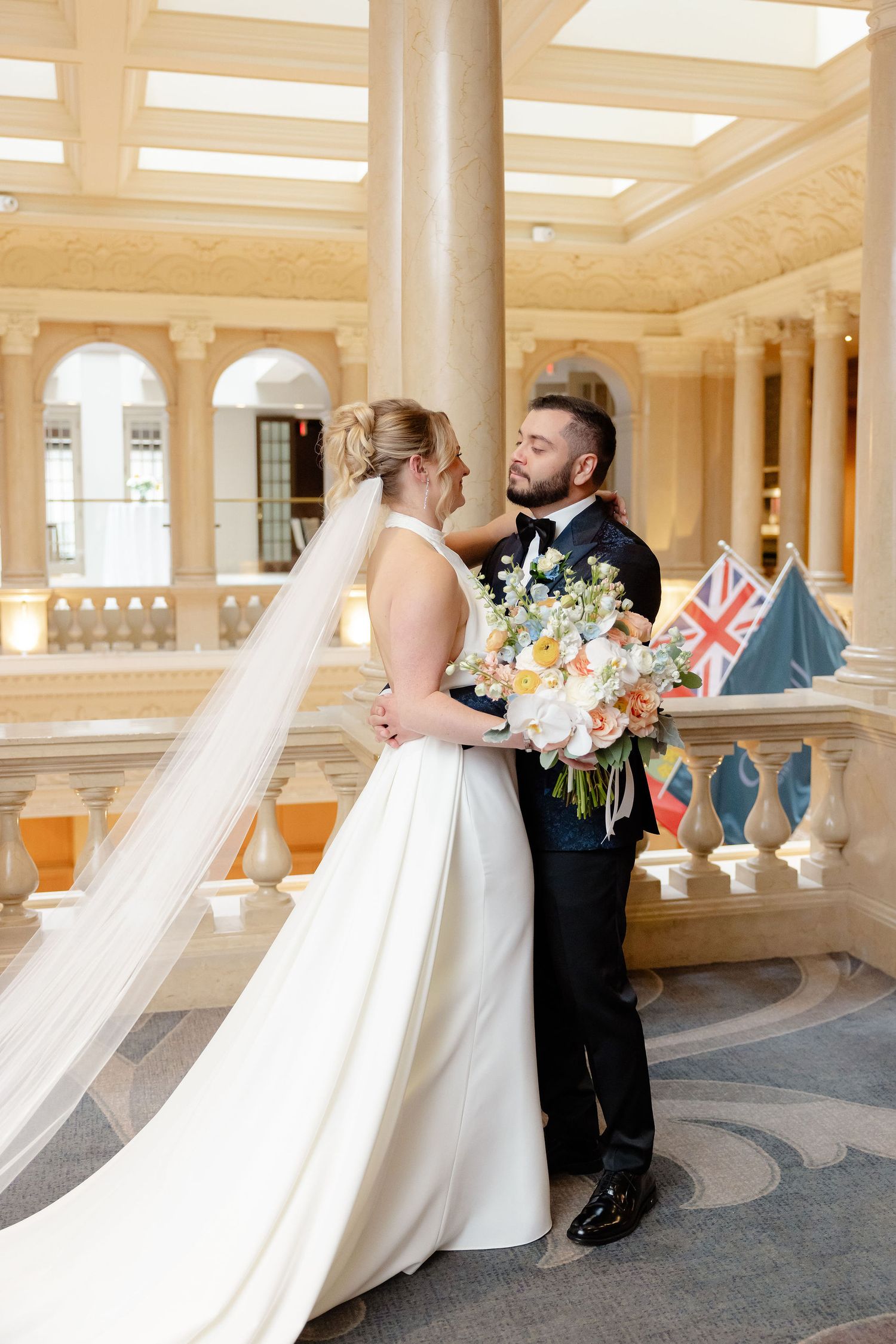 Romantic wedding moment in an ornate hallway with columns and flags, featuring a bride with flowing veil.