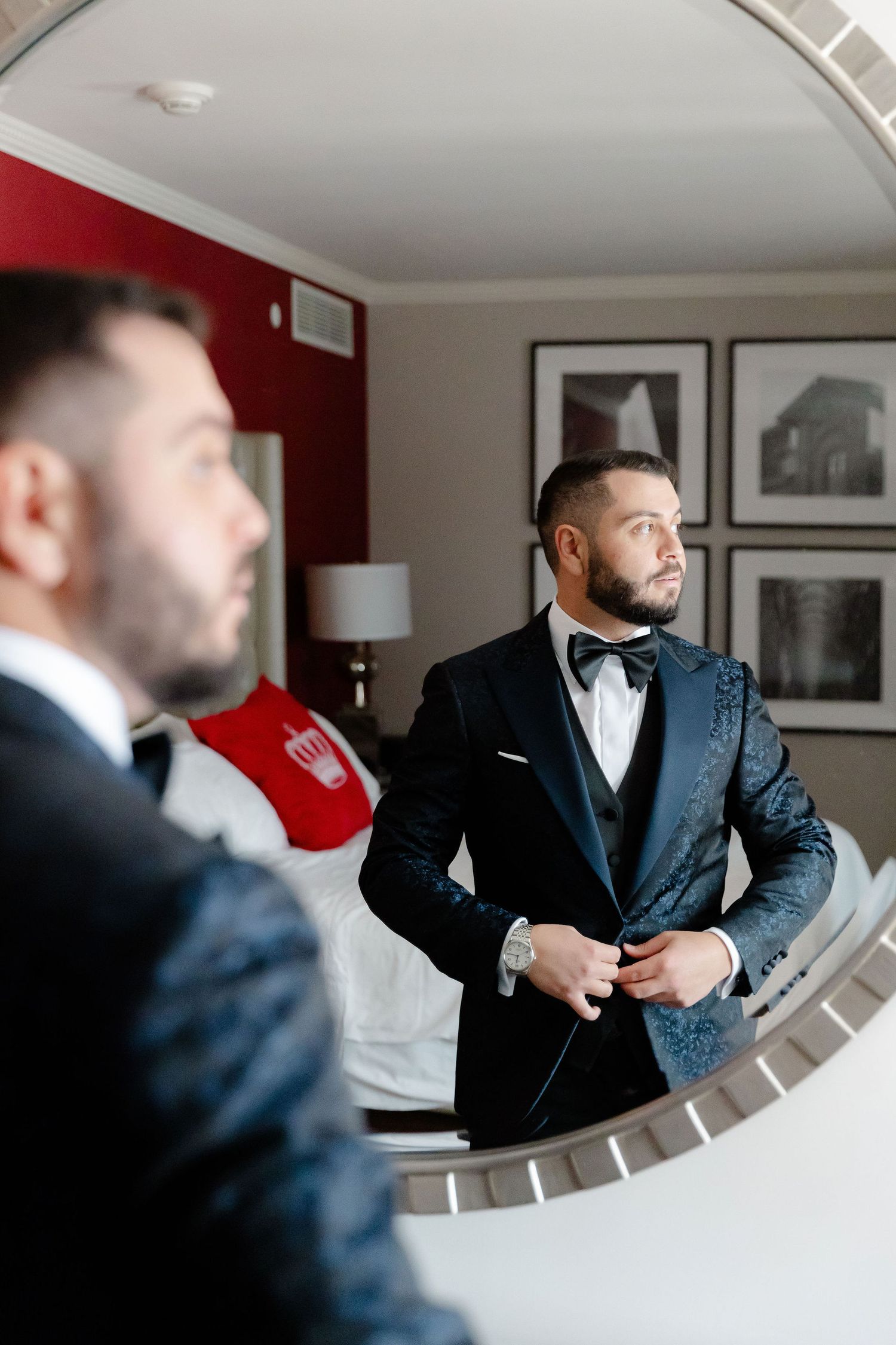 A reflection in a round mirror shows a groom adjusting his navy blue tuxedo jacket.