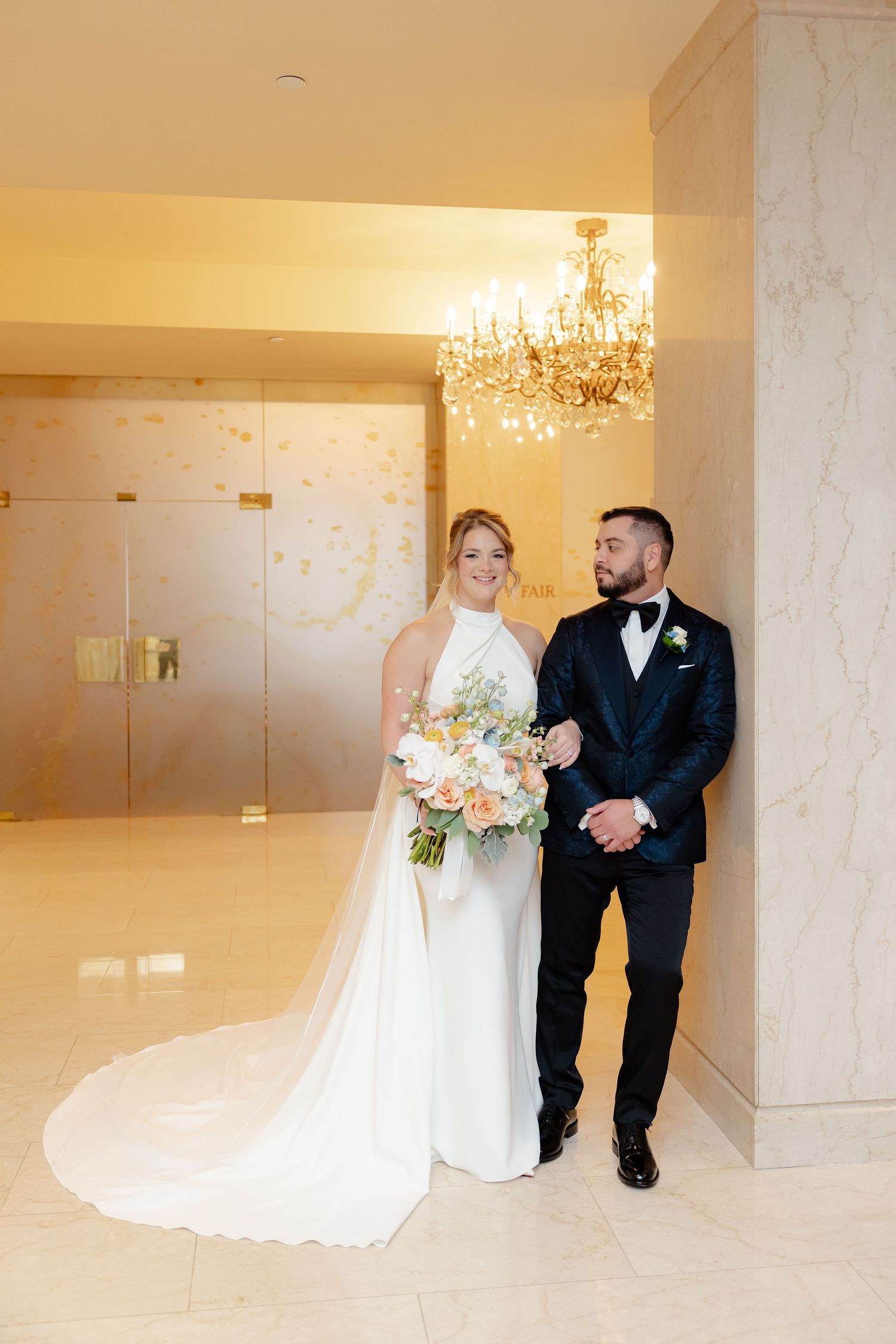 Wedding couple poses playfully in an elegant marble-walled venue with chandeliers.