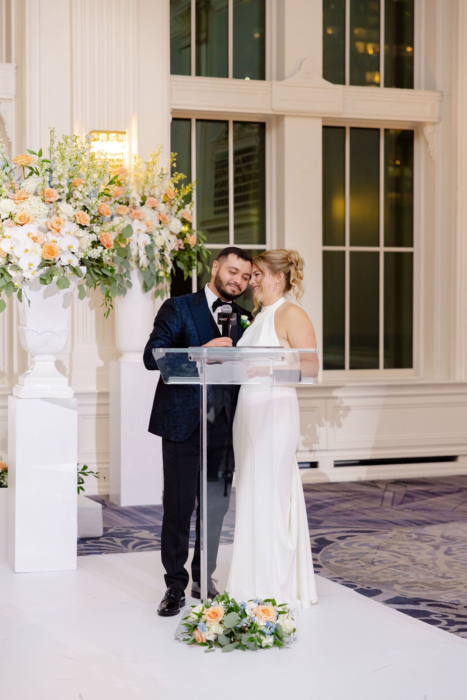 Wedding couple sharing a tender moment beside floral arrangements and white pedestals in an elegant venue.