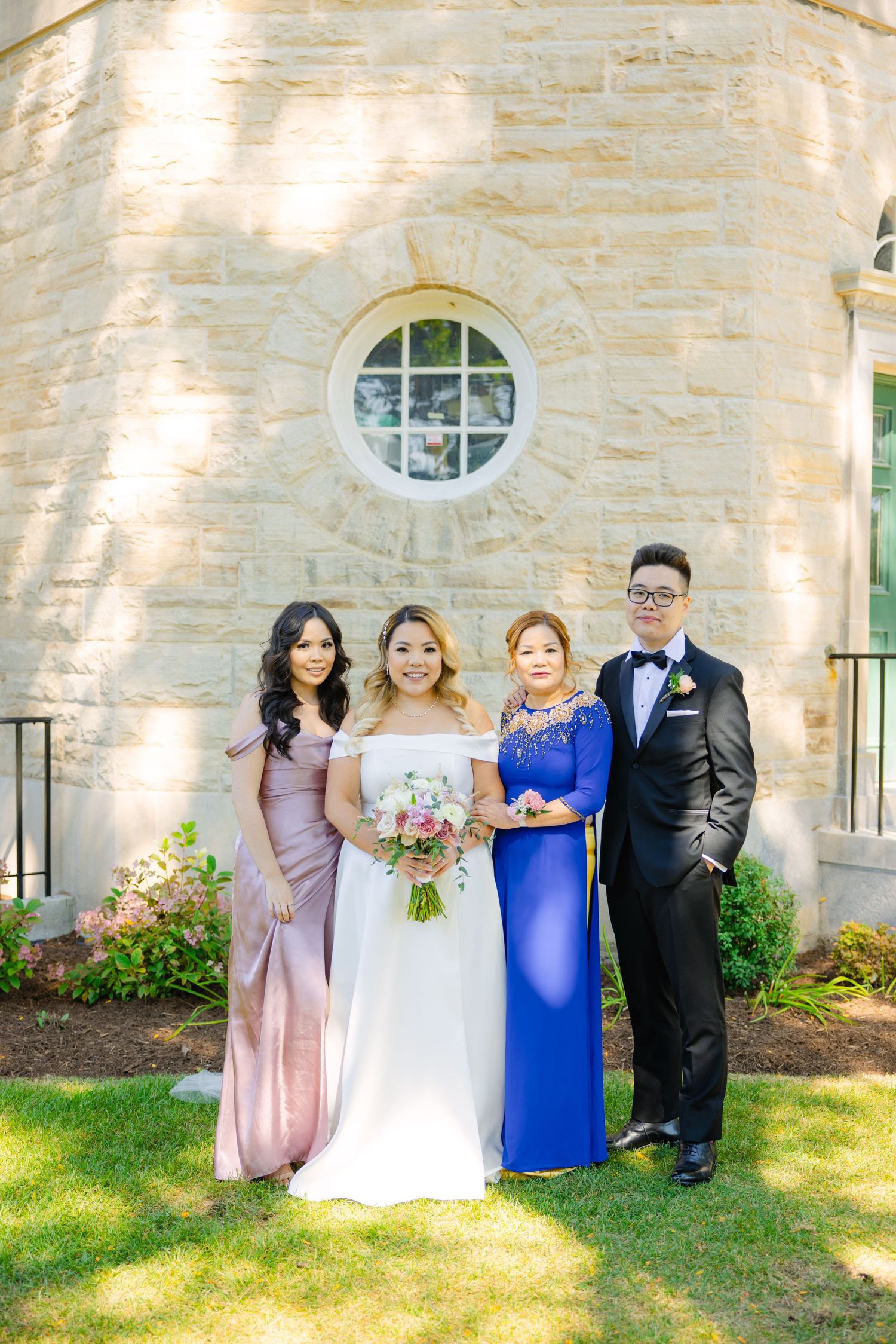 Wedding party poses outdoors by stone wall with round window wearing formal attire in pink, white and blue.