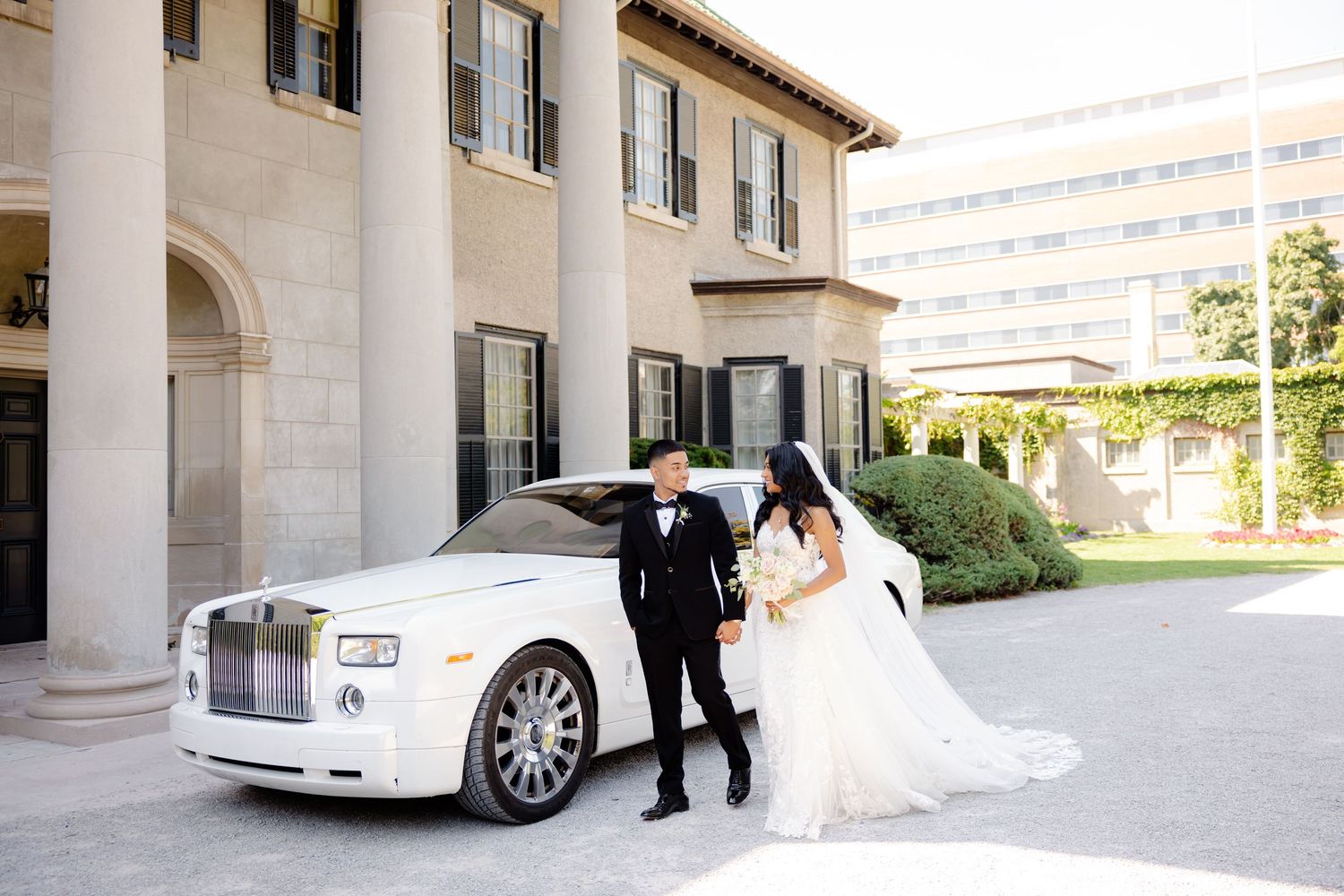 A wedding couple poses with their luxury Rolls-Royce outside an elegant Mediterranean-style building.