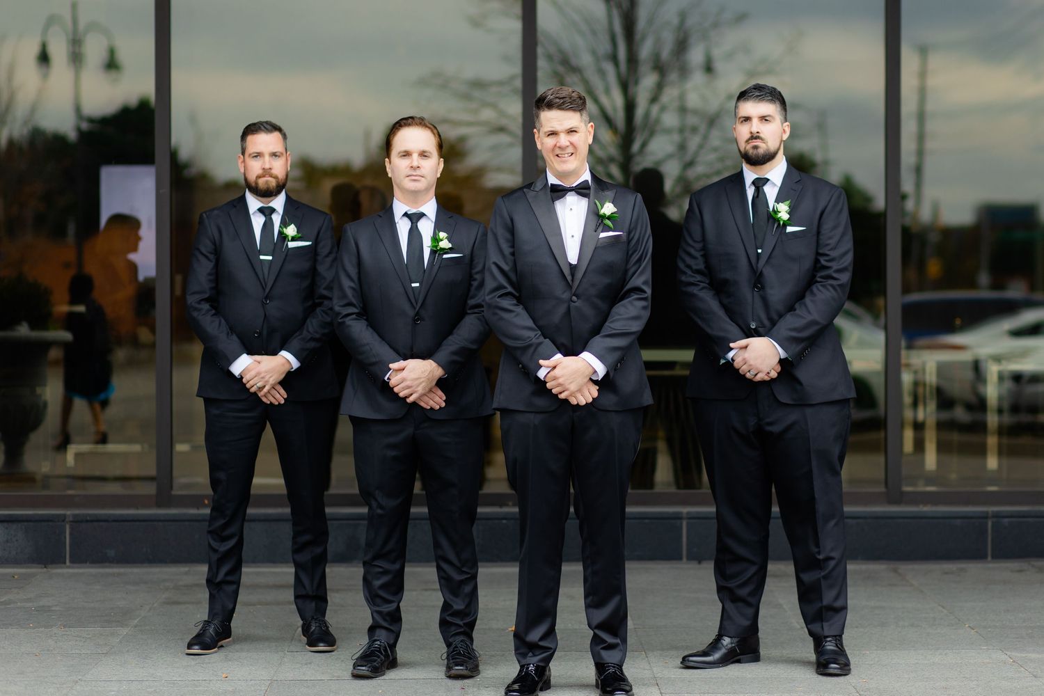 Four groomsmen in matching dark suits and boutonnieres stand formally outside modern building.