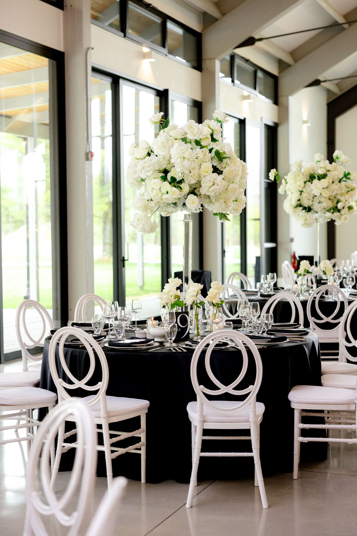 Modern wedding reception setup with white chairs, black tablecloths, and elevated white floral centerpieces.
