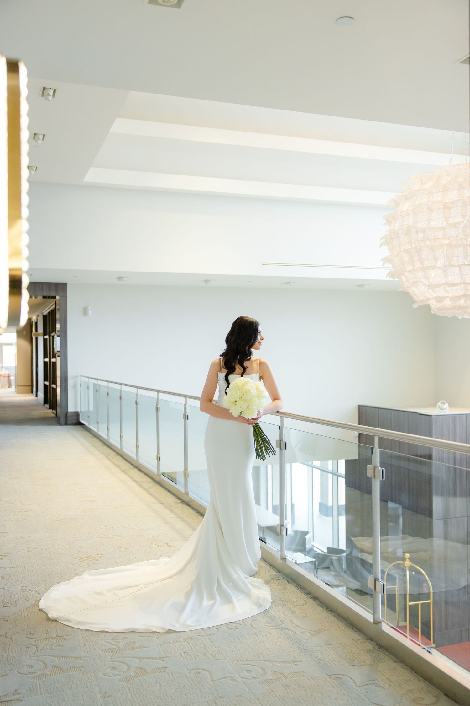 A bride in a fitted white wedding gown stands elegantly by a glass railing in a modern venue.