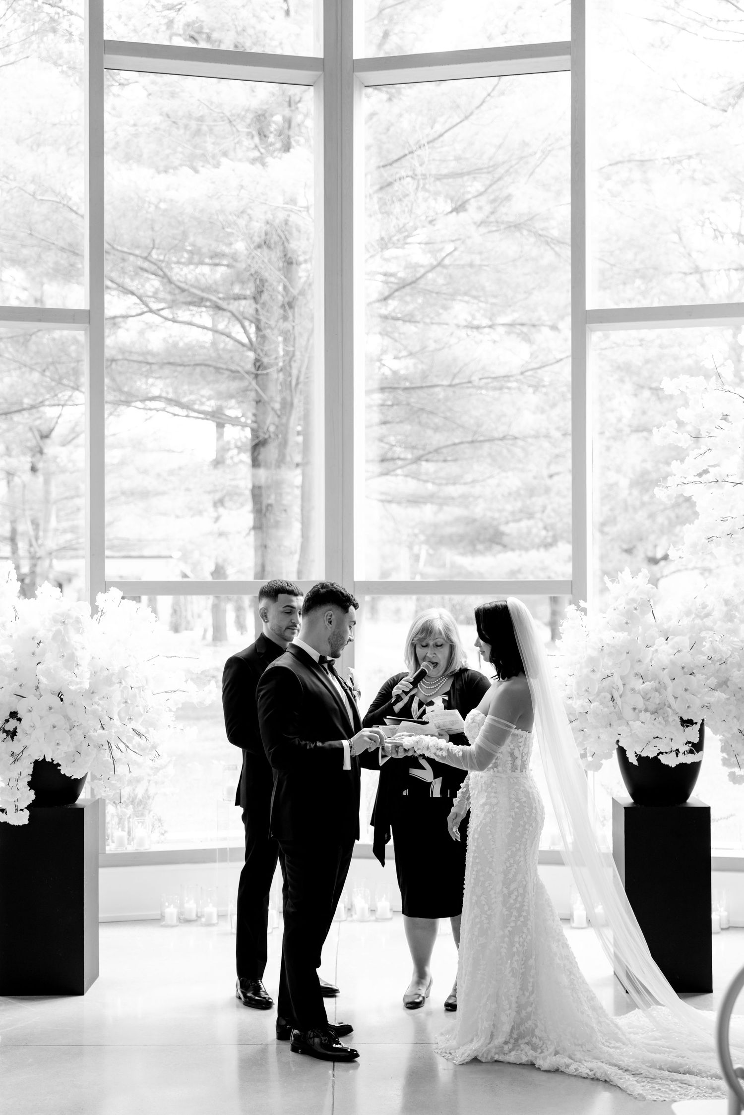 Black and white photo of wedding ceremony with tall windows and elegant floral decor.