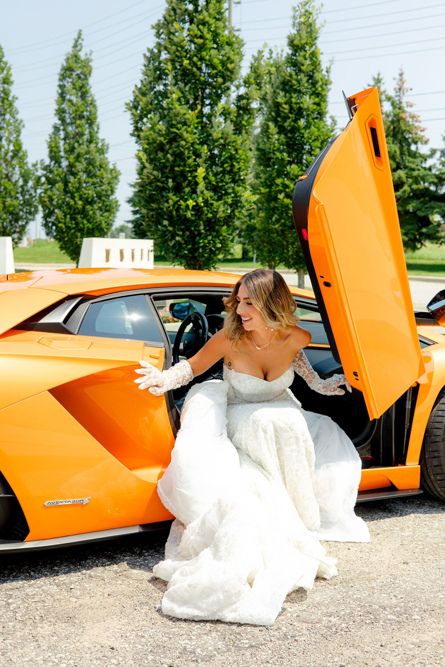A bride in a white wedding dress emerges from a bright orange luxury sports car with scissor doors.