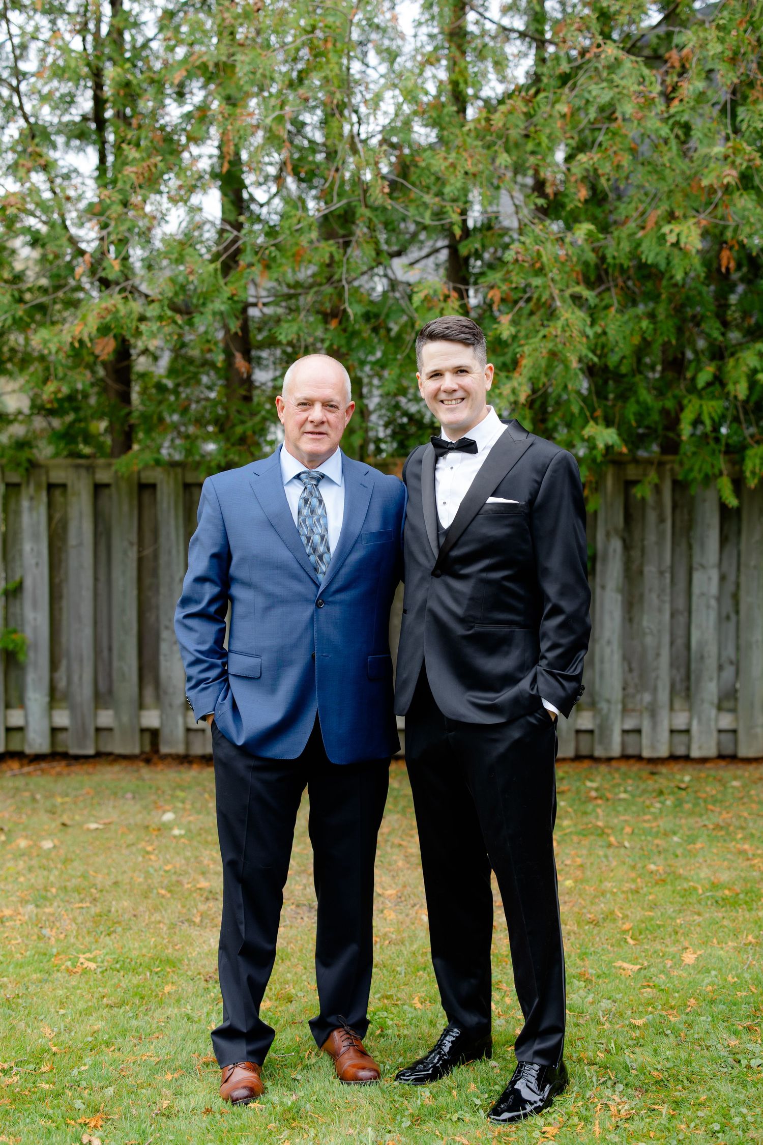 Two people in formal attire stand together against a backdrop of green trees.