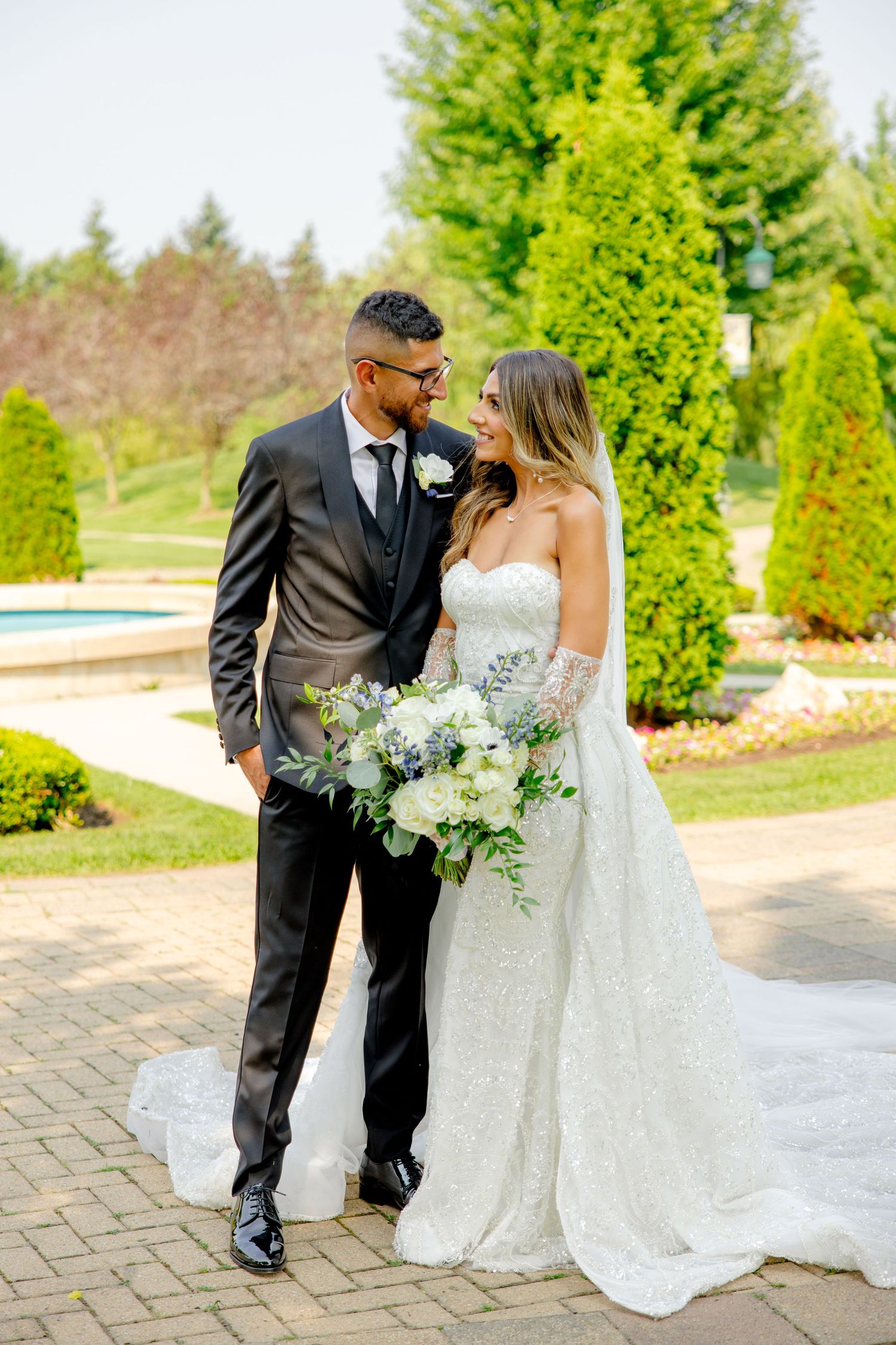 Newlyweds share an intimate moment in a garden with tall evergreen trees and a brick pathway.