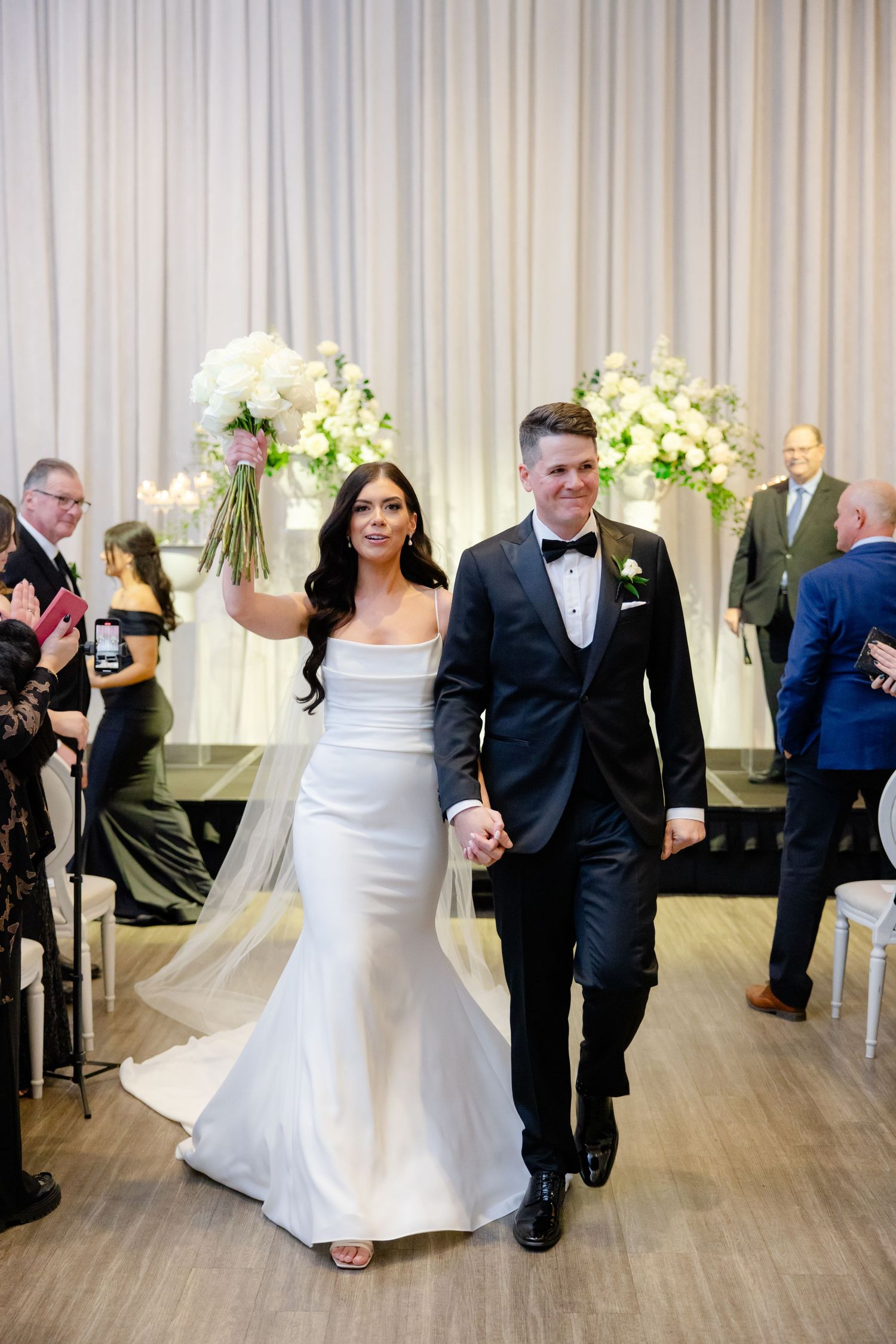 Newlyweds walking down aisle together after ceremony with white floral decorations in background.