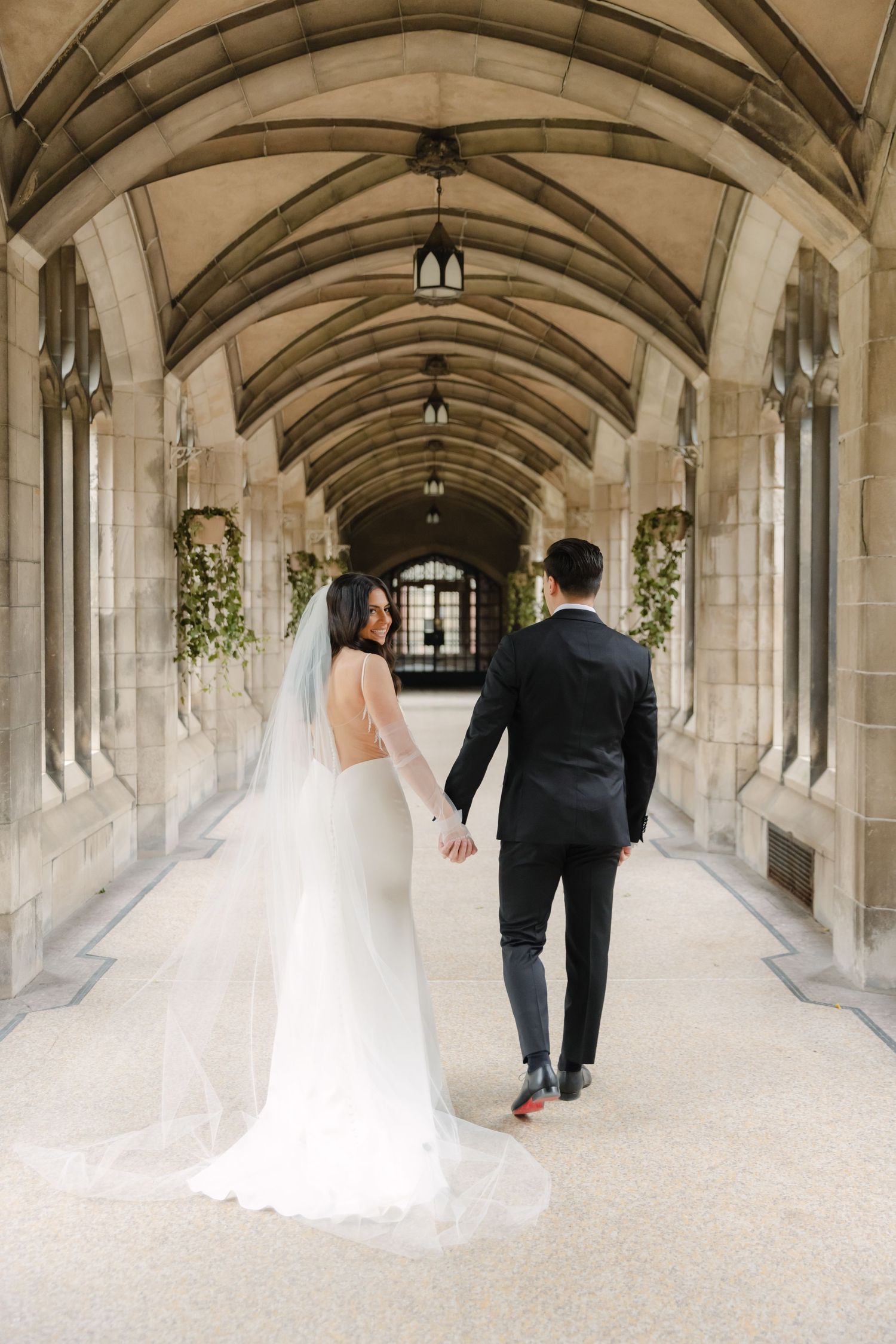Wedding couple walks through elegant stone archway corridor decorated with hanging greenery and lanterns.