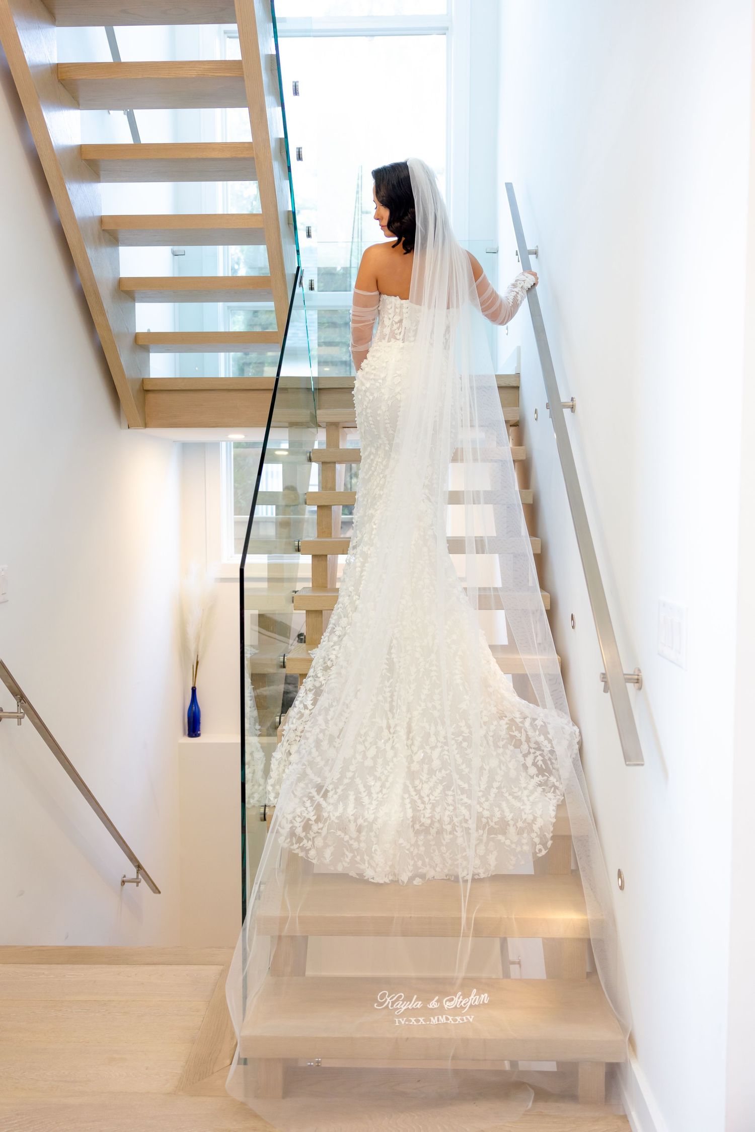 A bride in a lace wedding gown ascends modern wooden stairs with glass railings.
