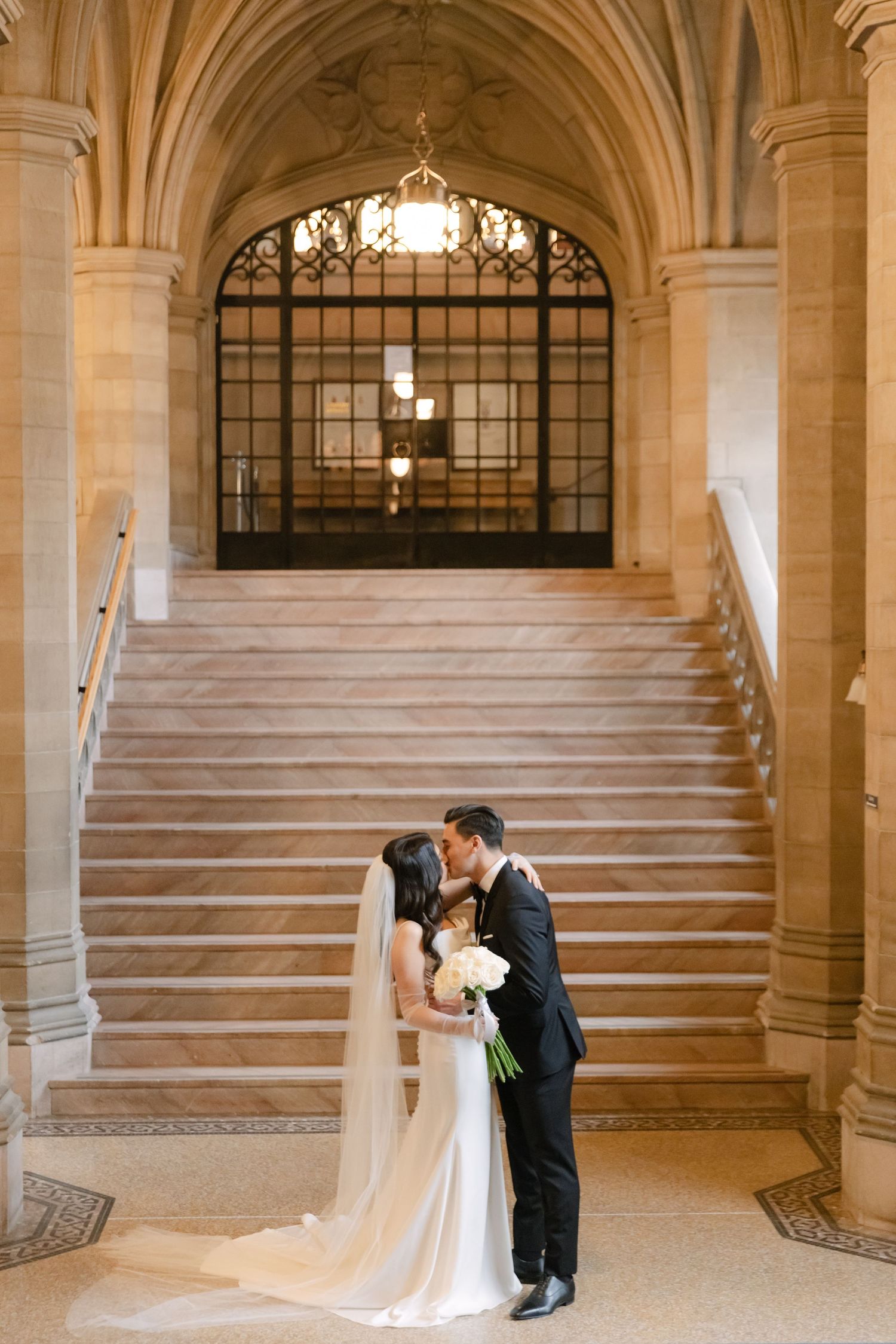 A romantic wedding photograph taken at the base of a grand architectural staircase with arched details.