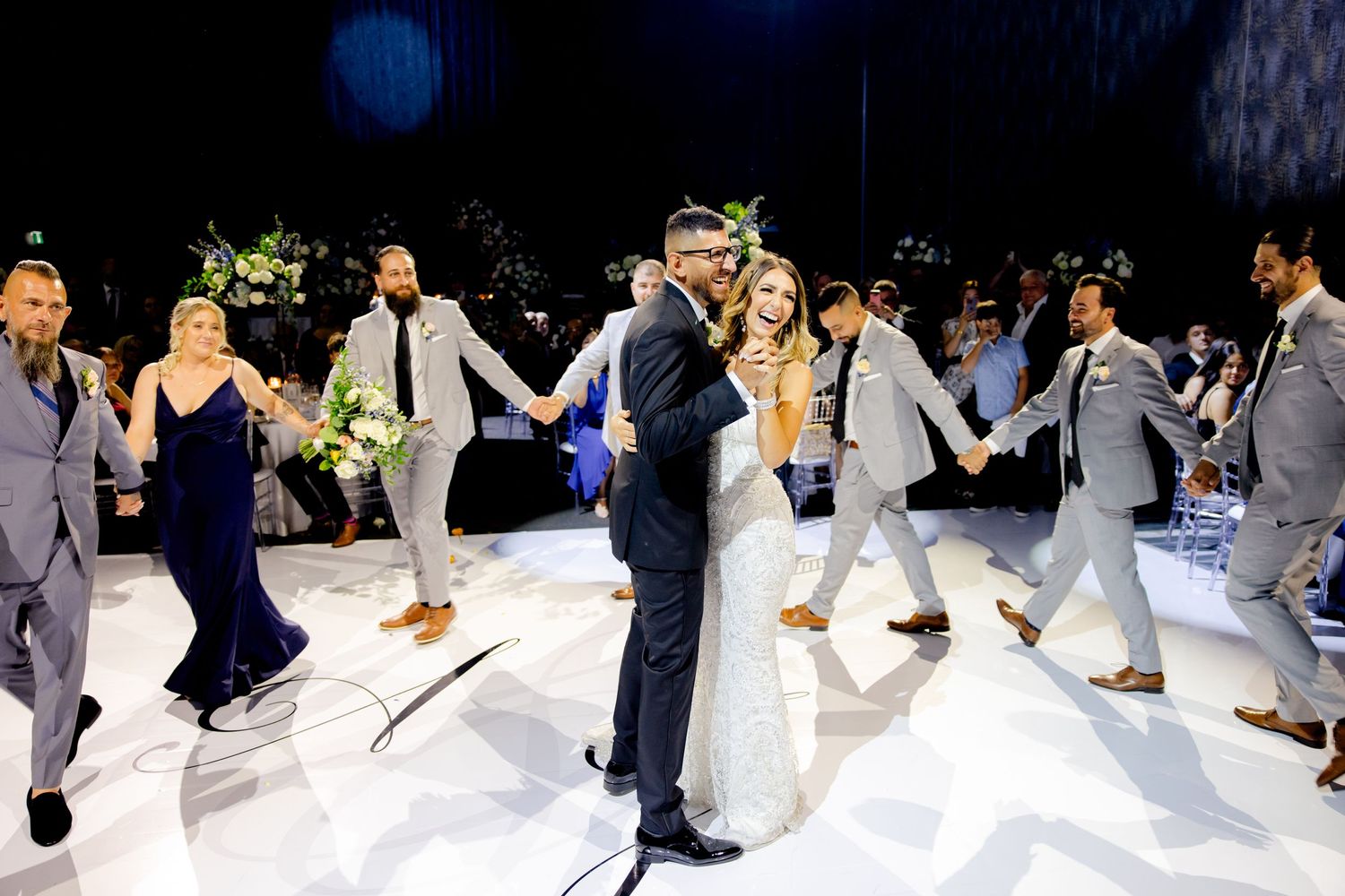 Wedding party forms a circle around dancing couple on illuminated white dance floor.