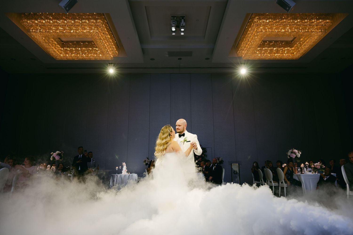Wedding couple dances surrounded by dramatic fog effects and elegant chandelier lighting.
