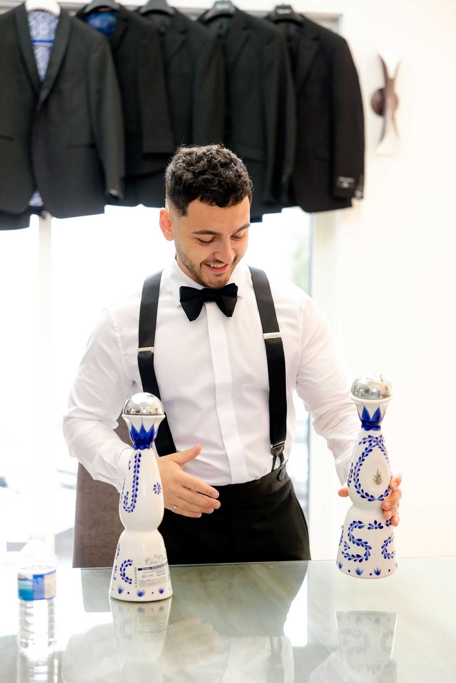 A person in formal attire with suspenders and bow tie stands by decorative blue and white bottles.