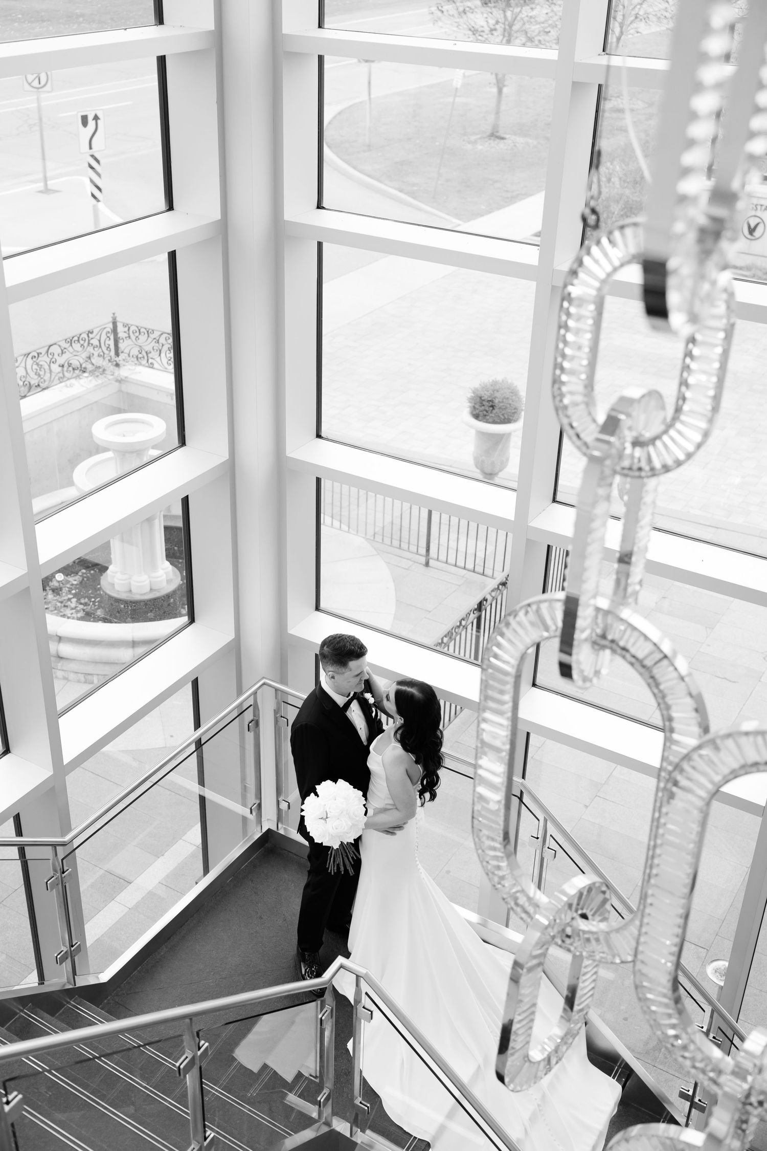 Black and white photograph of a wedding couple in an elegant stairwell with natural light streaming through windows.