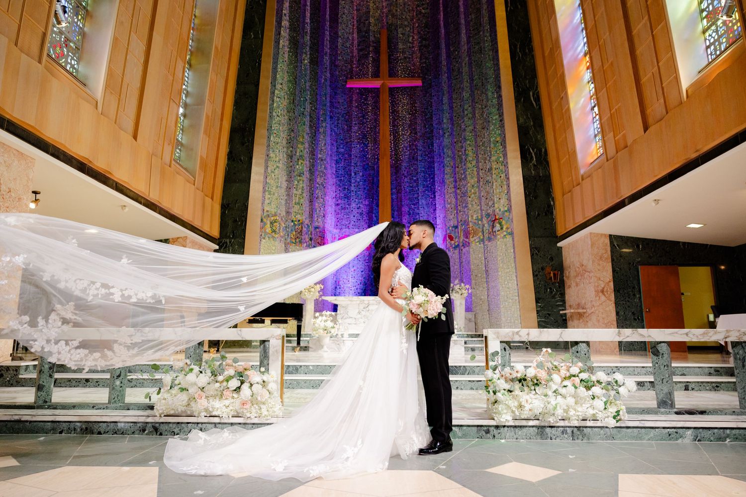 Wedding couple embraces in front of purple-lit church altar with flowing cathedral veil.