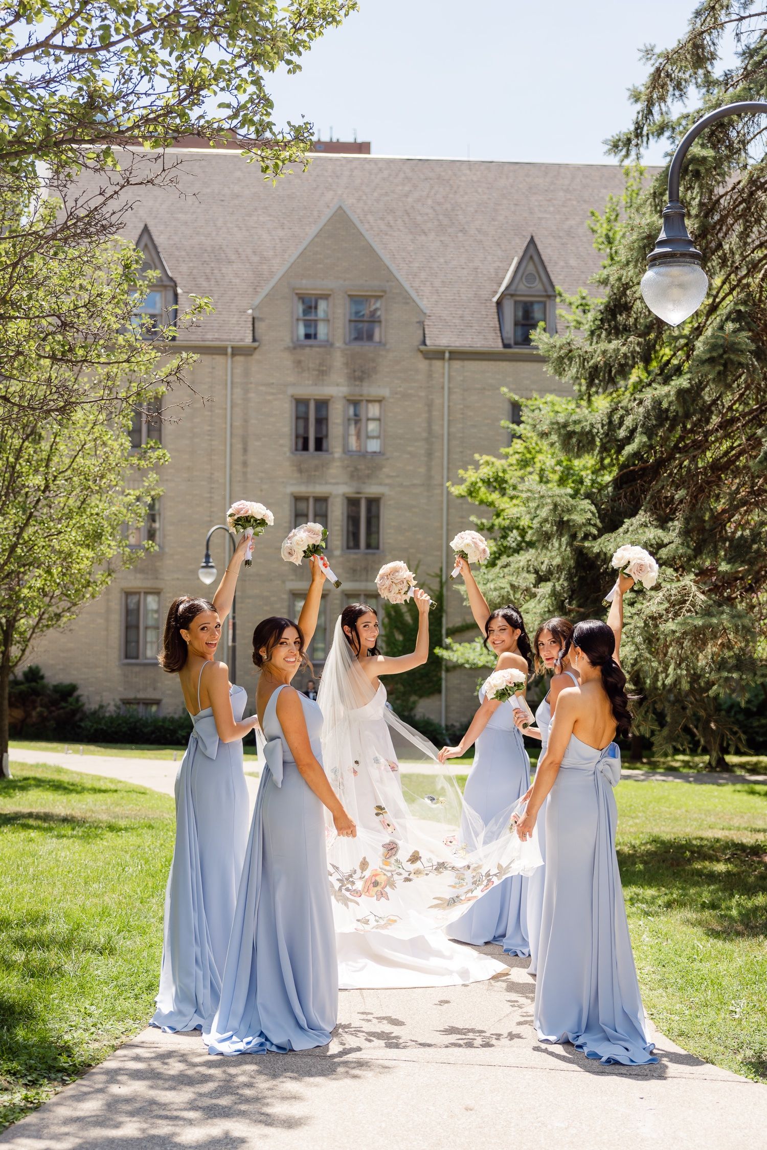 Bridesmaids in blue dresses lift bouquets while holding bride's veil in sunlit garden.