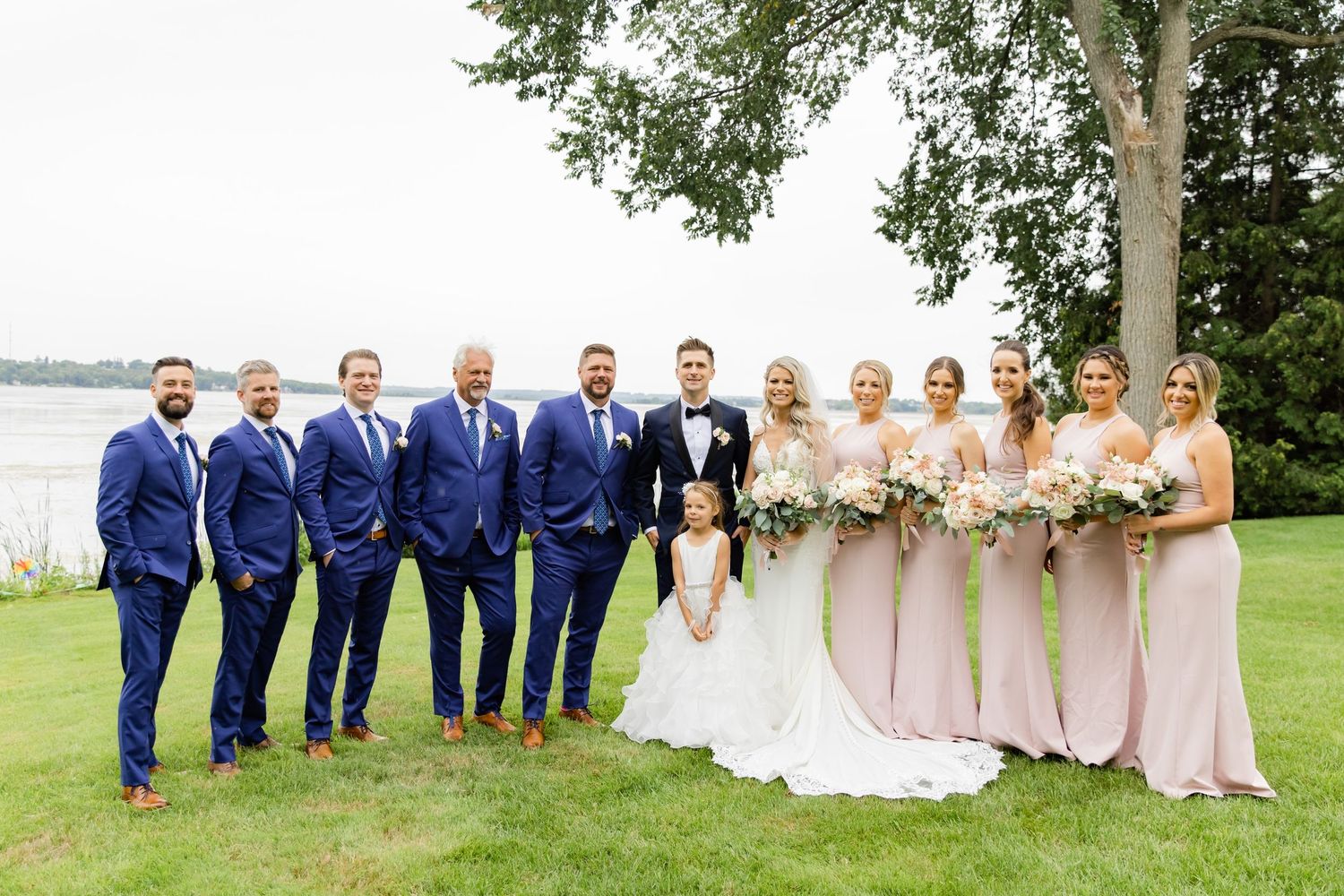 Wedding party in blue suits and pink dresses poses by waterfront.