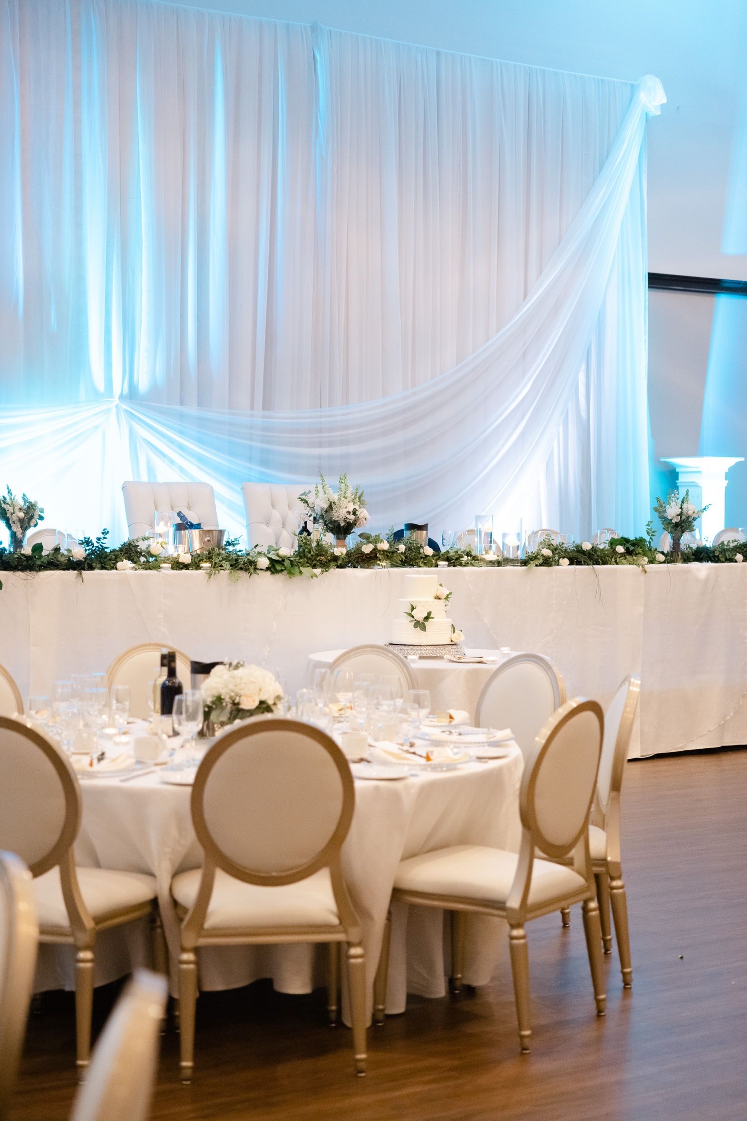 Modern wedding reception setup with white draping, floral arrangements and cream Louis XVI style chairs.