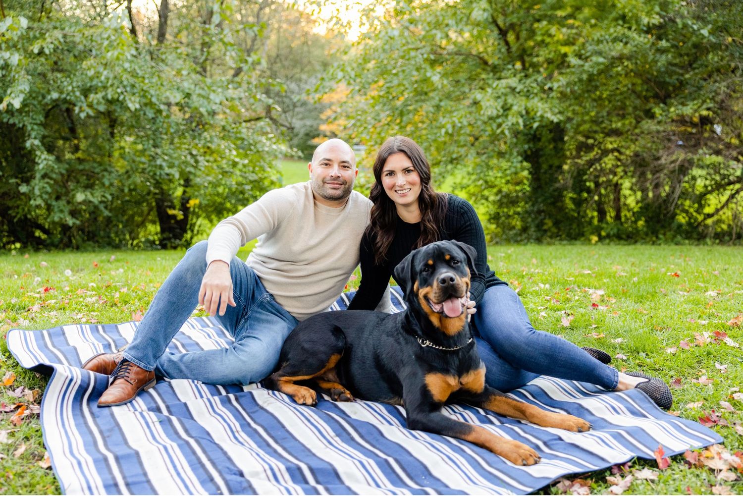 Couple relaxing on striped blanket with their Rottweiler dog in autumn park setting.