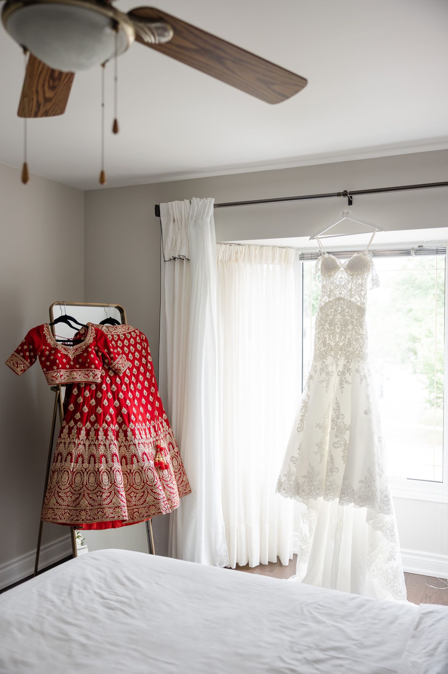 Red traditional dress and white wedding gown displayed in bedroom with ceiling fan.